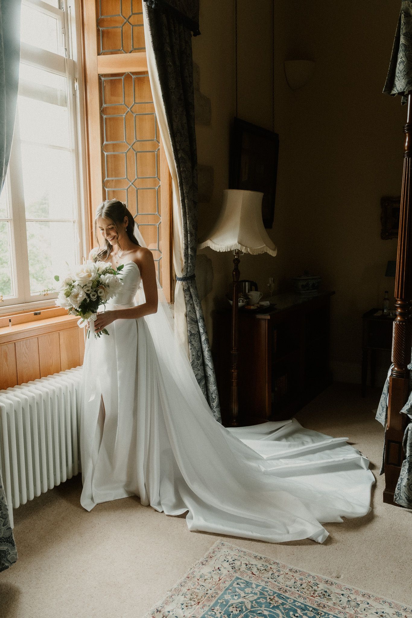 Bride in elegant white gown with long train holds bouquet by sunlit leaded window in vintage room.
