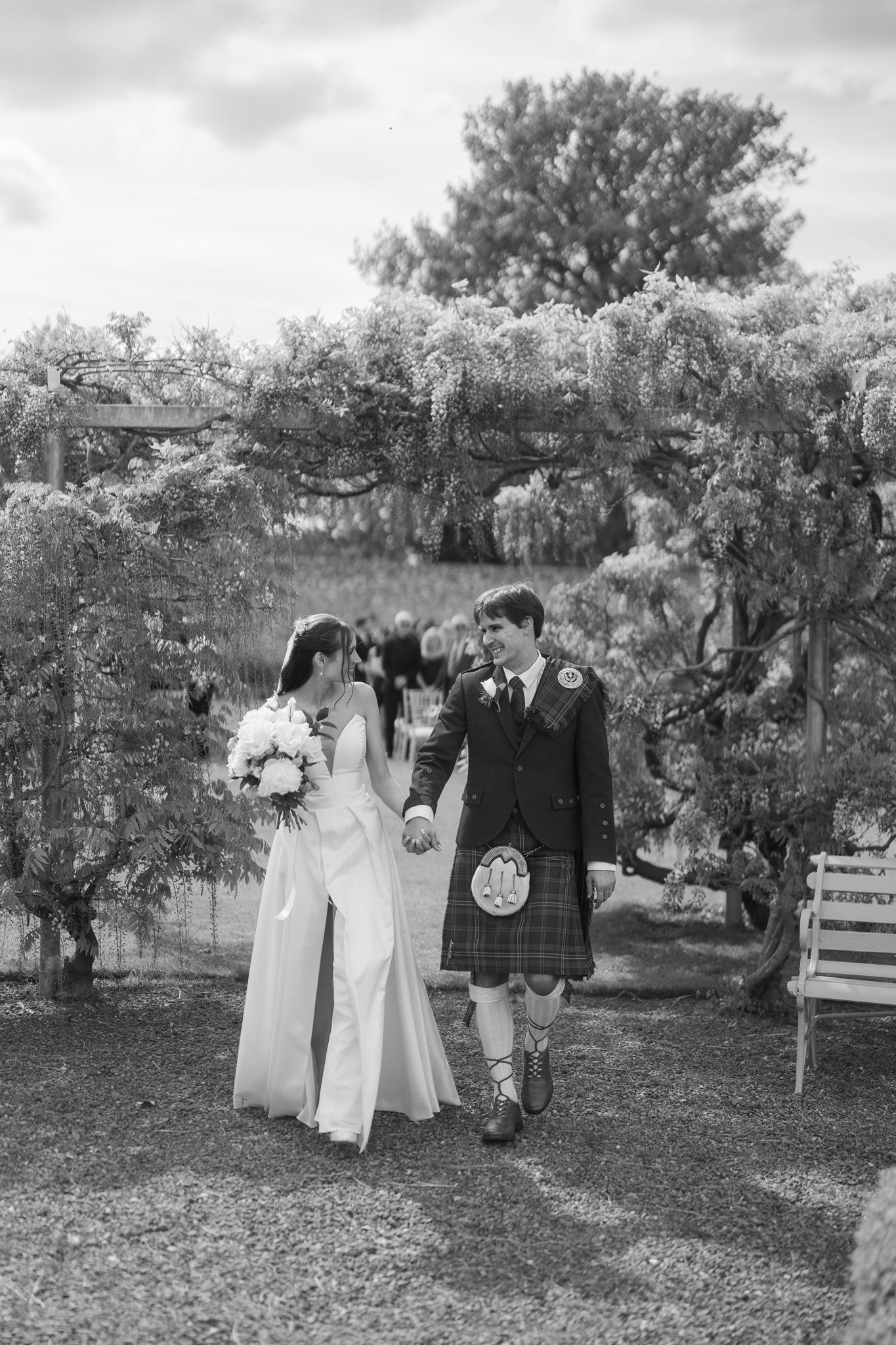 Black and white photo of a bride and groom in a kilt walking hand in hand under a floral arch in a garden.