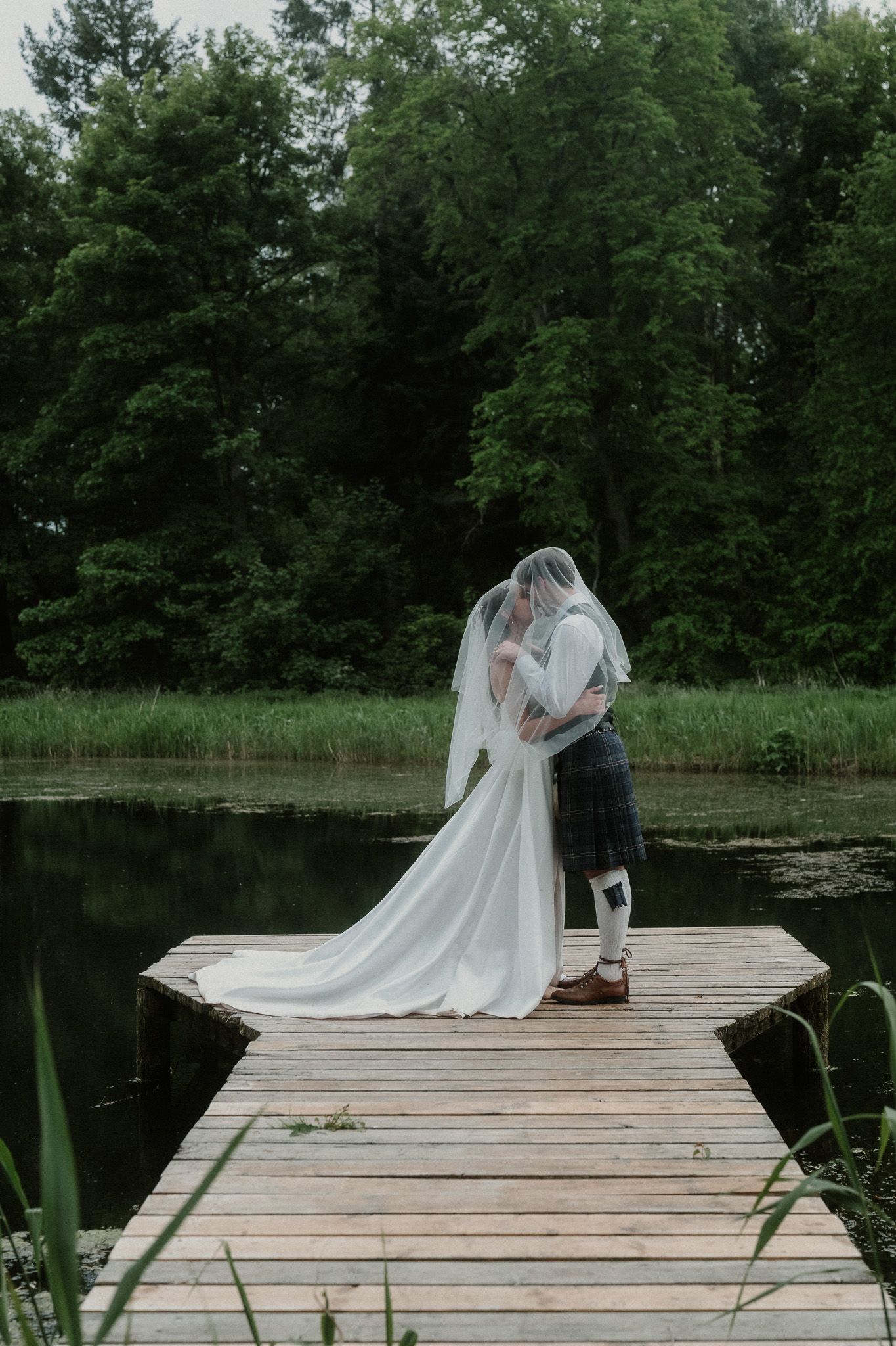 Bride and groom sharing a kiss under a veil on a wooden dock by a serene lake surrounded by lush green trees.
