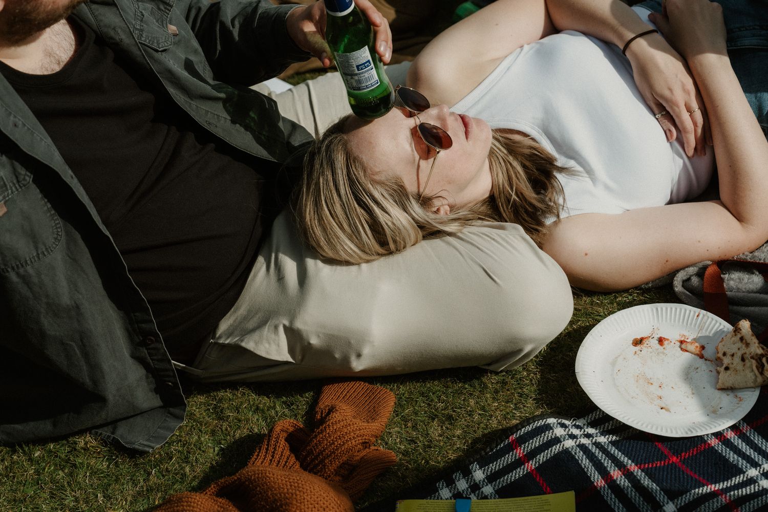 Woman in sunglasses rests her head on a man's lap on a picnic blanket while he holds a green bottle, with a plate of food nearby.