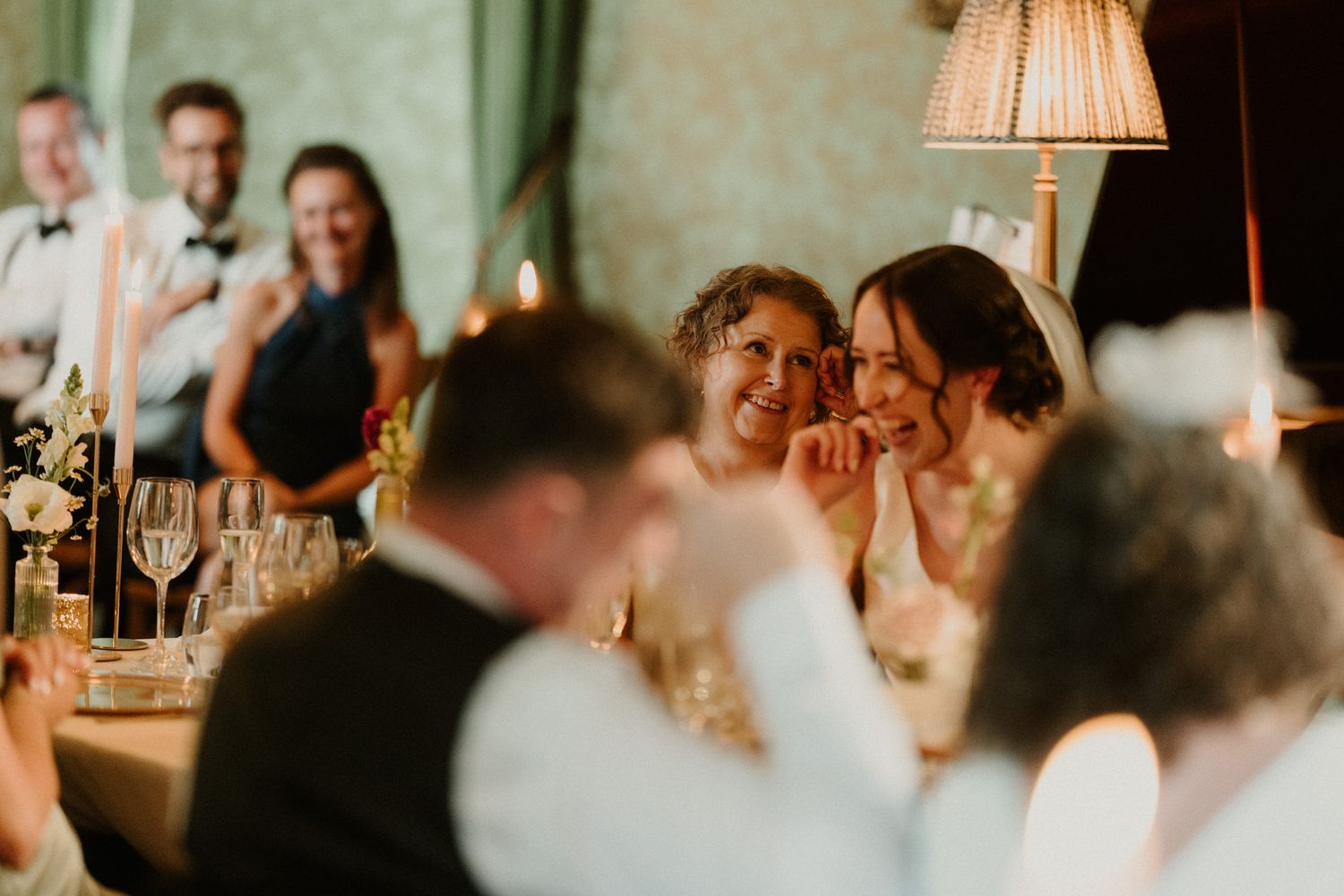 Smiling wedding guests laugh together at a candlelit reception table with wine glasses, flowers, and a warm lamp glowing in the background.