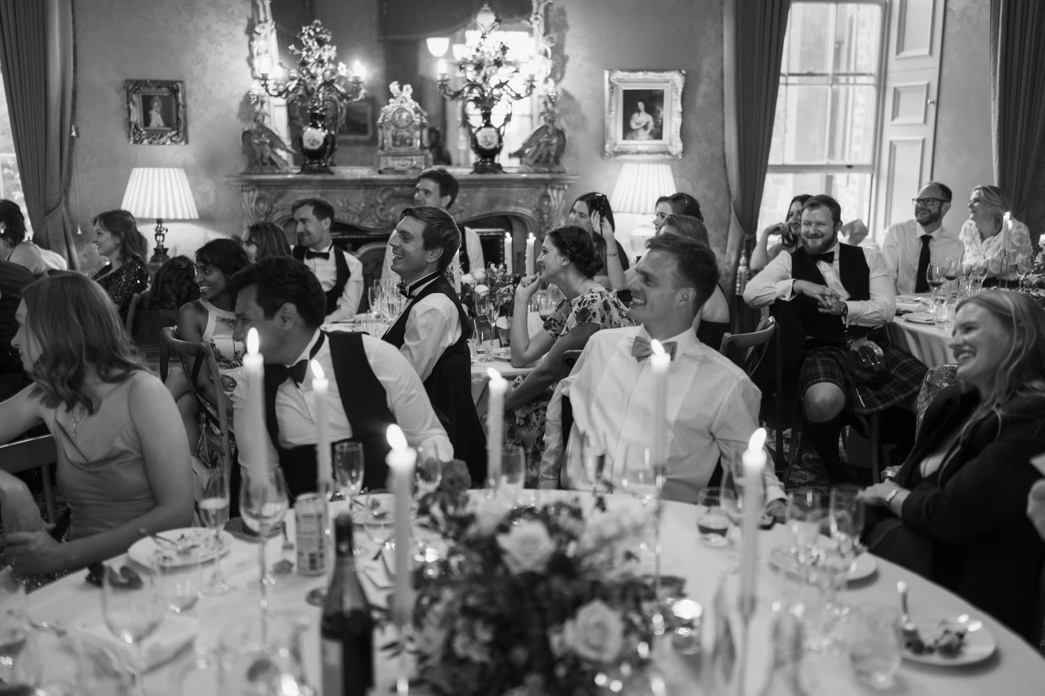 Wedding guests laugh and celebrate around a candlelit banquet table with flowers and glassware in an elegant black and white reception hall.