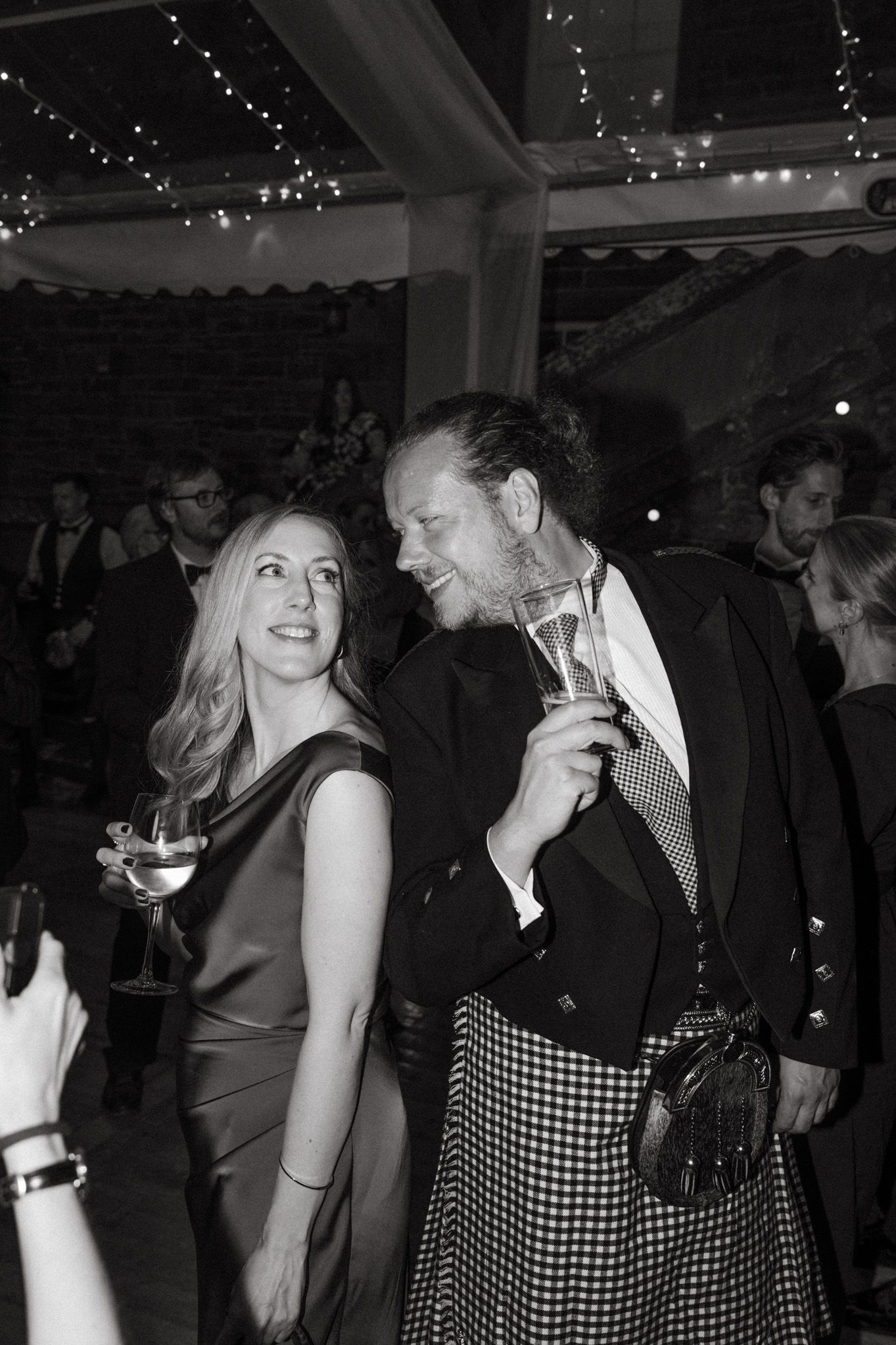 Black and white photo of a smiling woman in a satin dress and a man in a kilt holding drinks while dancing at a festive evening party.
