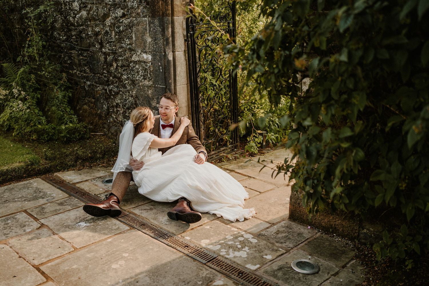 Bride and groom sitting together on stone steps surrounded by lush greenery at an outdoor wedding venue.