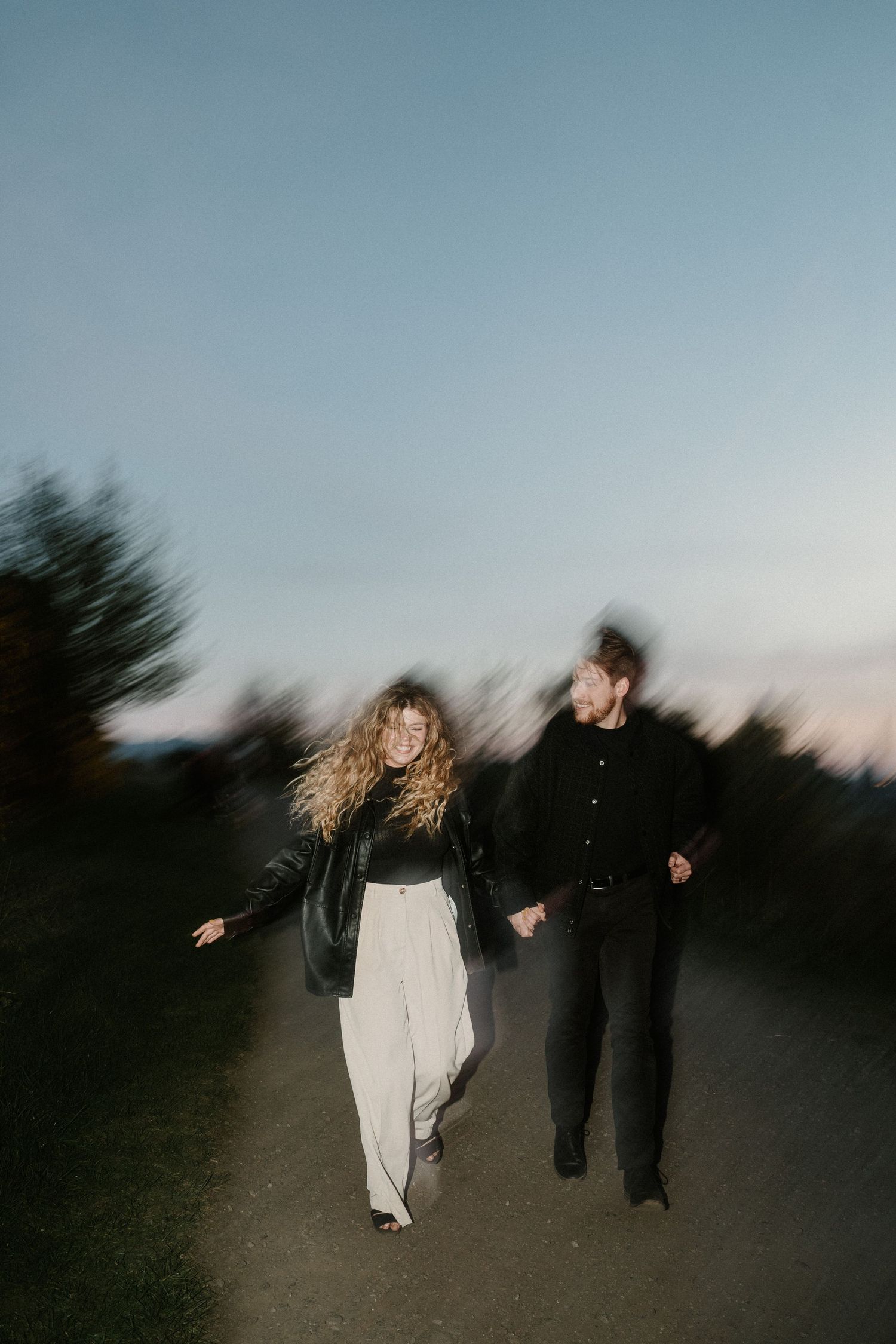Couple holding hands walking on a path at dusk, motion blur effect, woman in white pants and black jacket.