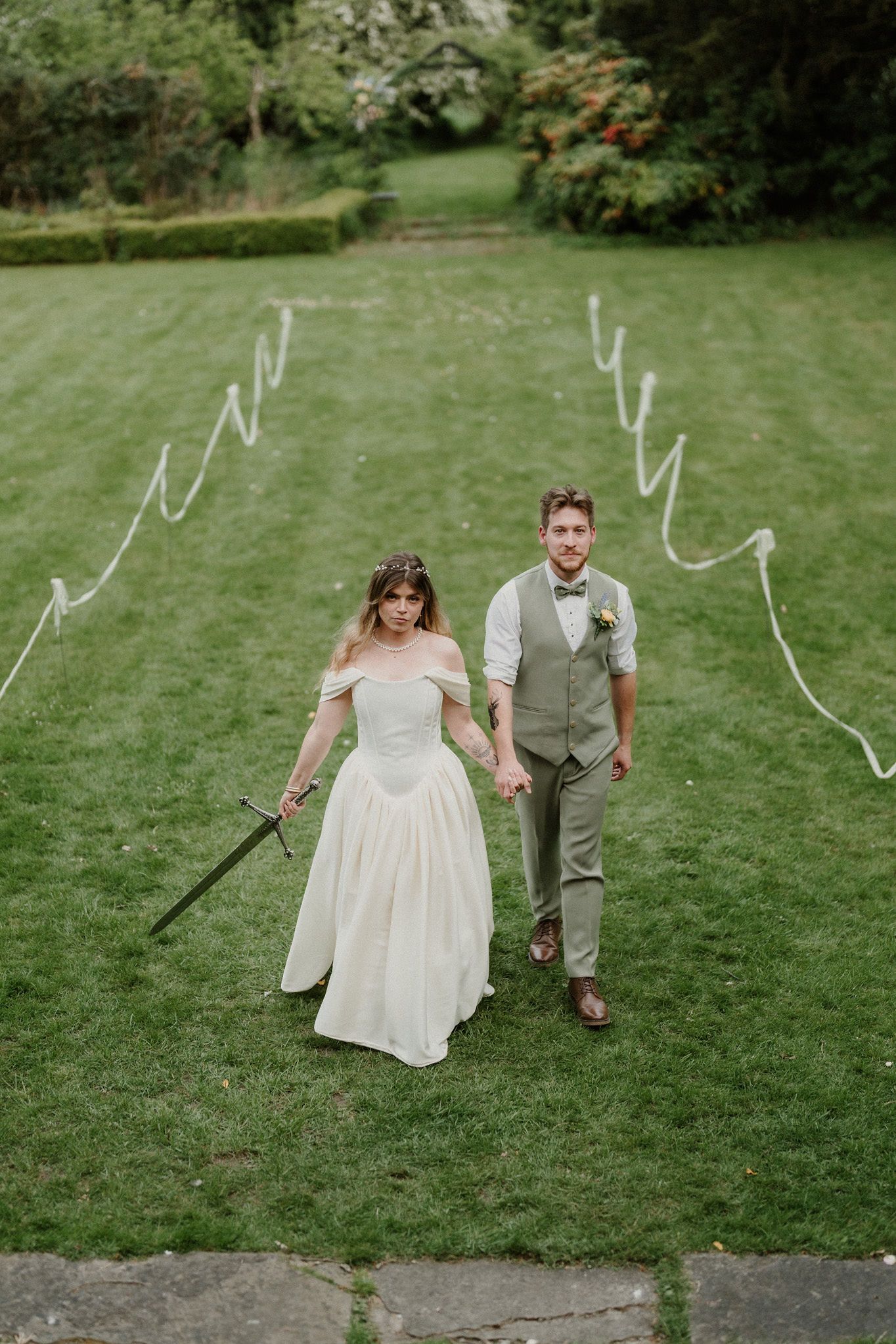 Bride in white gown holding sword and groom in green suit walking down garden path at wedding.