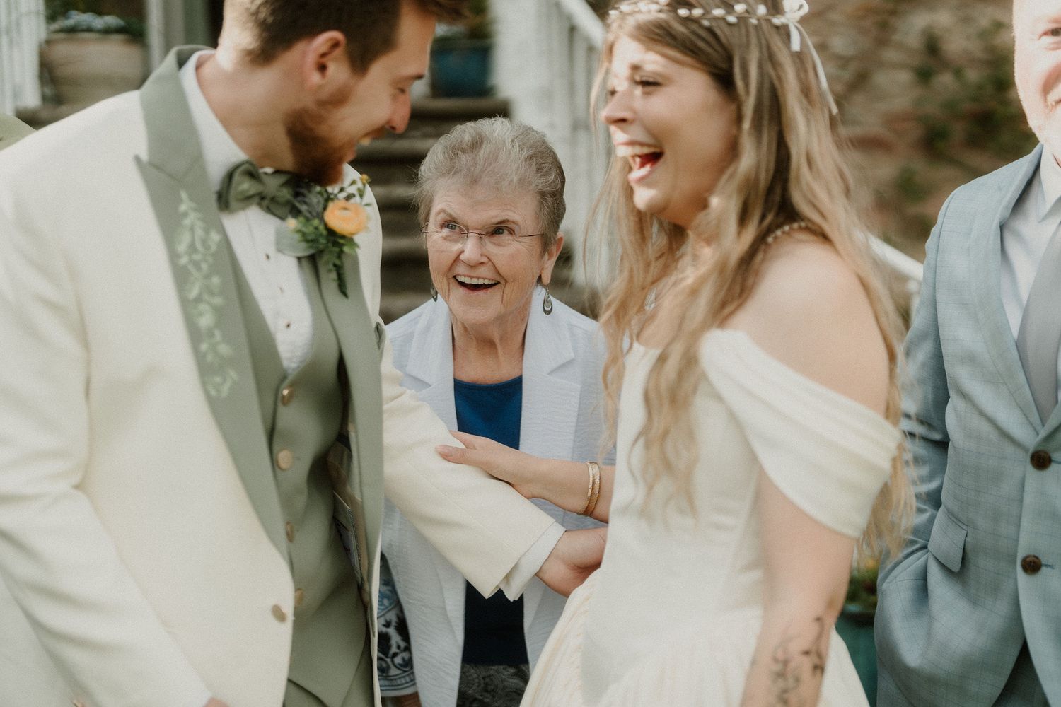 Elderly grandmother laughing joyfully with bride and groom in white wedding attire during outdoor wedding celebration.