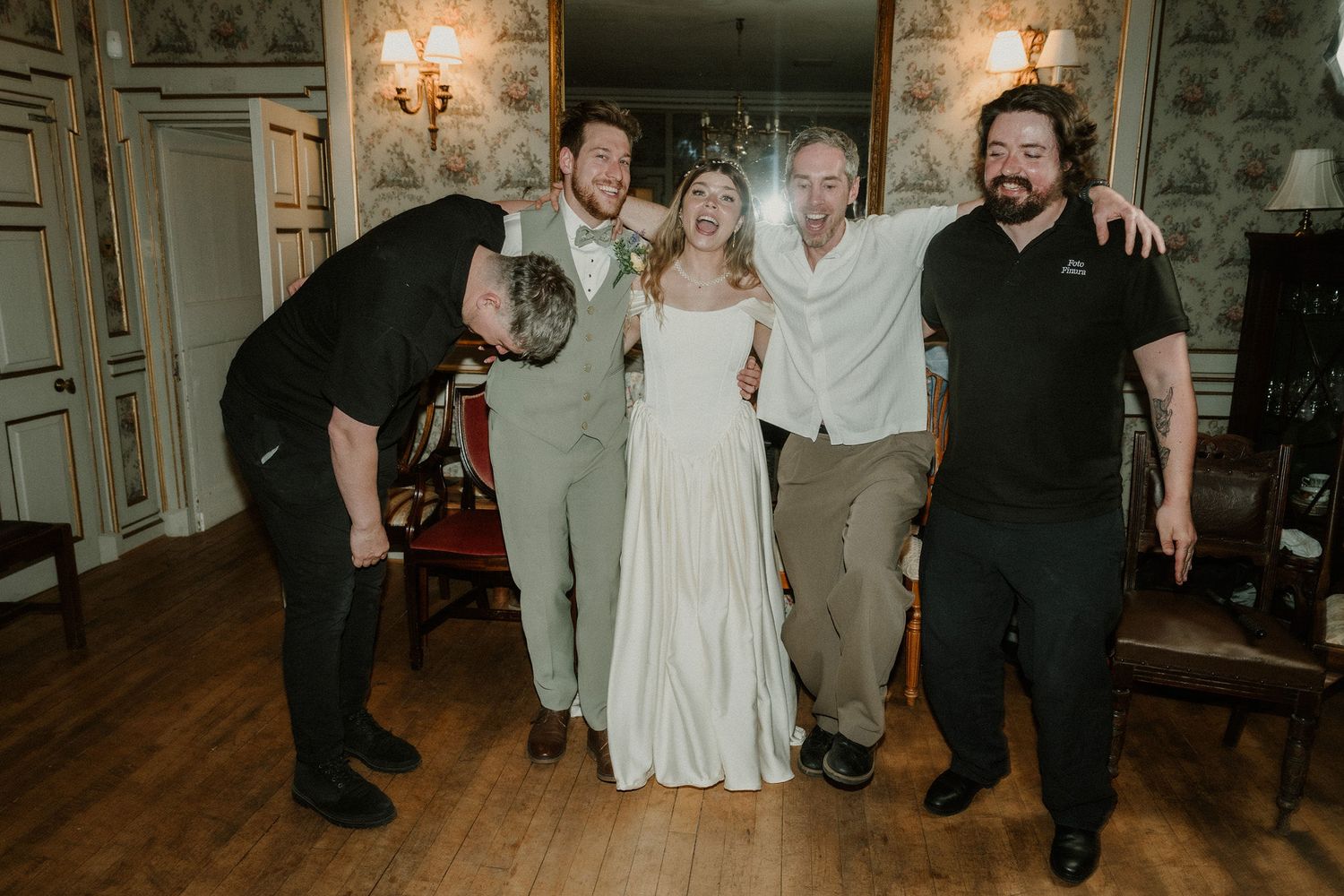 Bride in white gown poses joyfully with four guests in elegant wallpapered room at wedding reception.