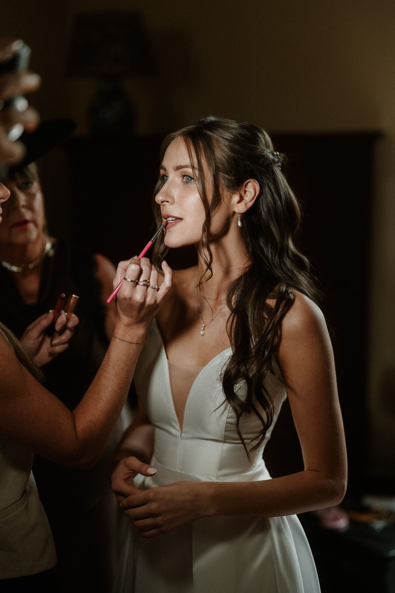 Bride in white gown gets lipstick applied by makeup artist during wedding day preparations.