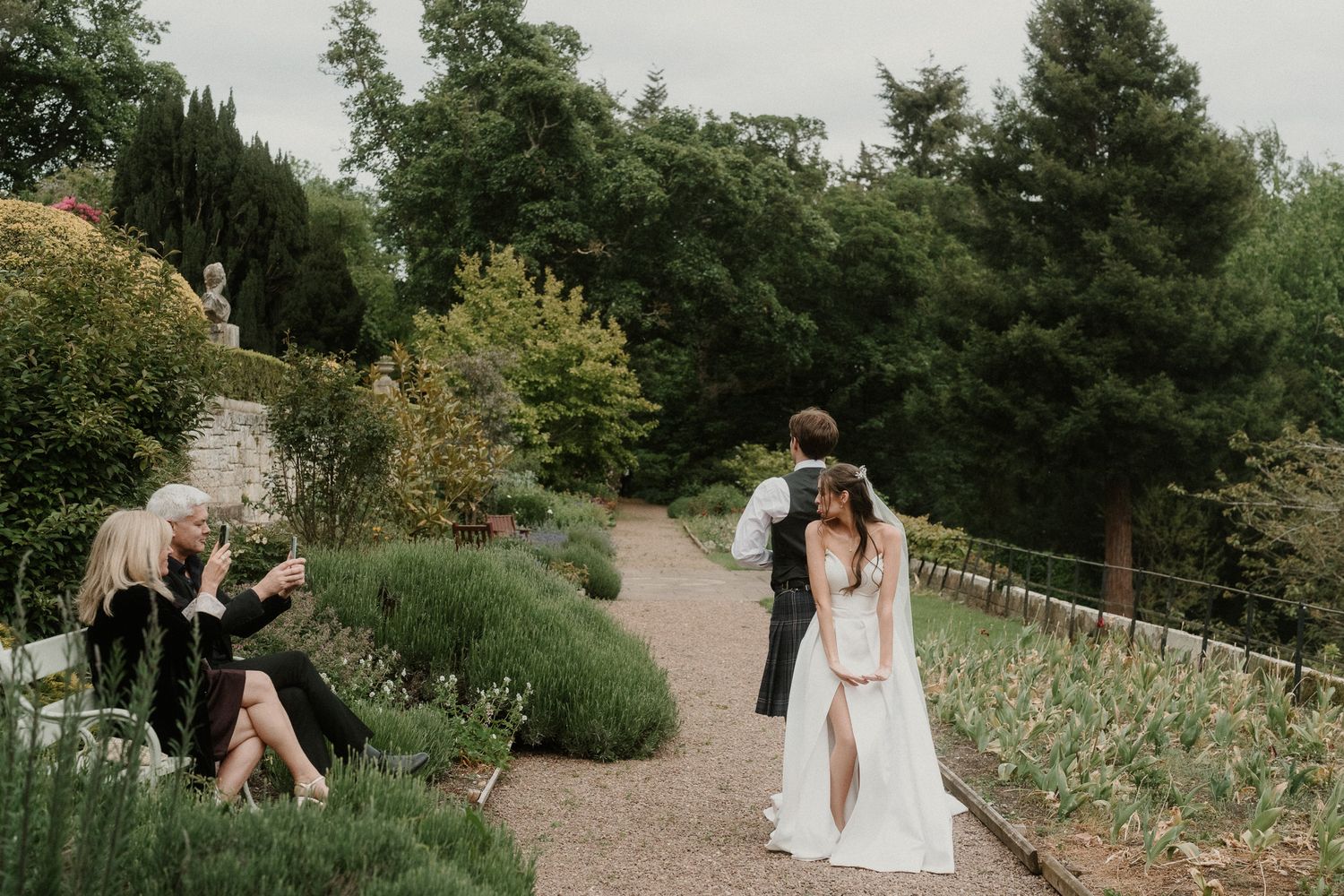 A bride and groom share a romantic moment in a lush garden, while guests relax nearby on a wedding day.