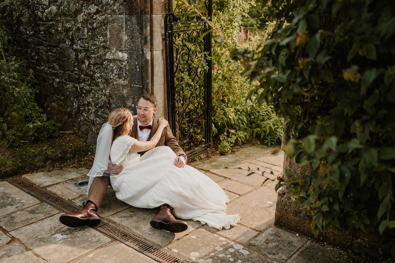 A romantic couple sits together on stone steps surrounded by lush greenery in a garden setting.