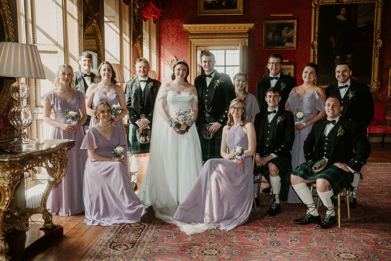 Wedding party seated in ornate historic room with red walls and elegant decor.