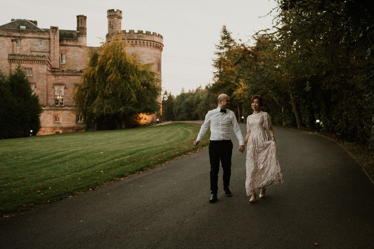 A couple strolls down a winding path surrounded by autumn trees near a stone building.