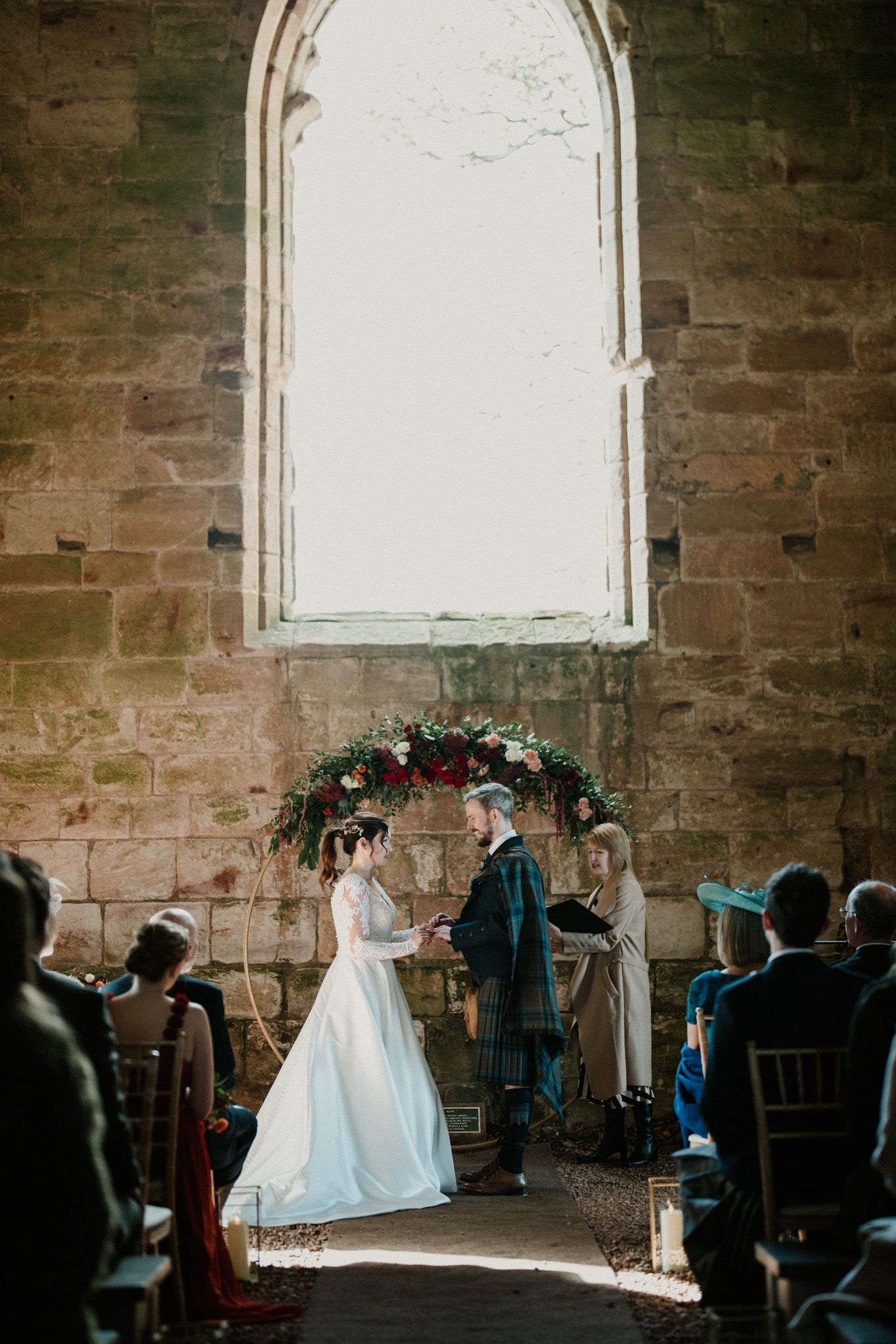 Wedding ceremony under an arched floral arrangement in a rustic stone church with gothic windows.