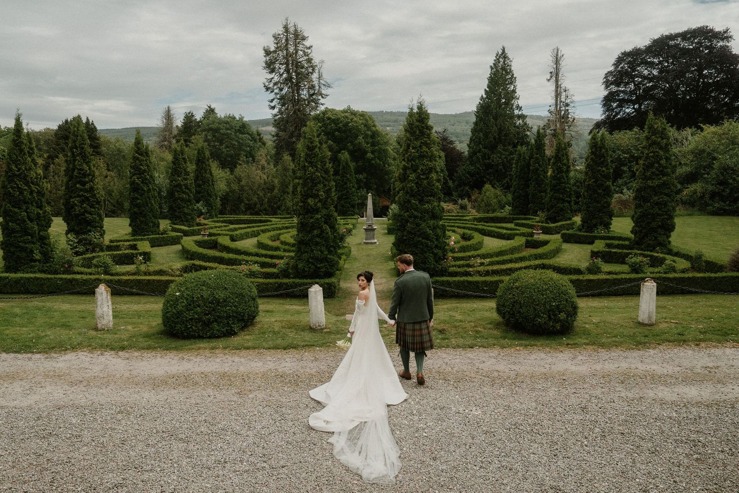 A couple walks through a manicured garden with topiary and hedges on an overcast day.