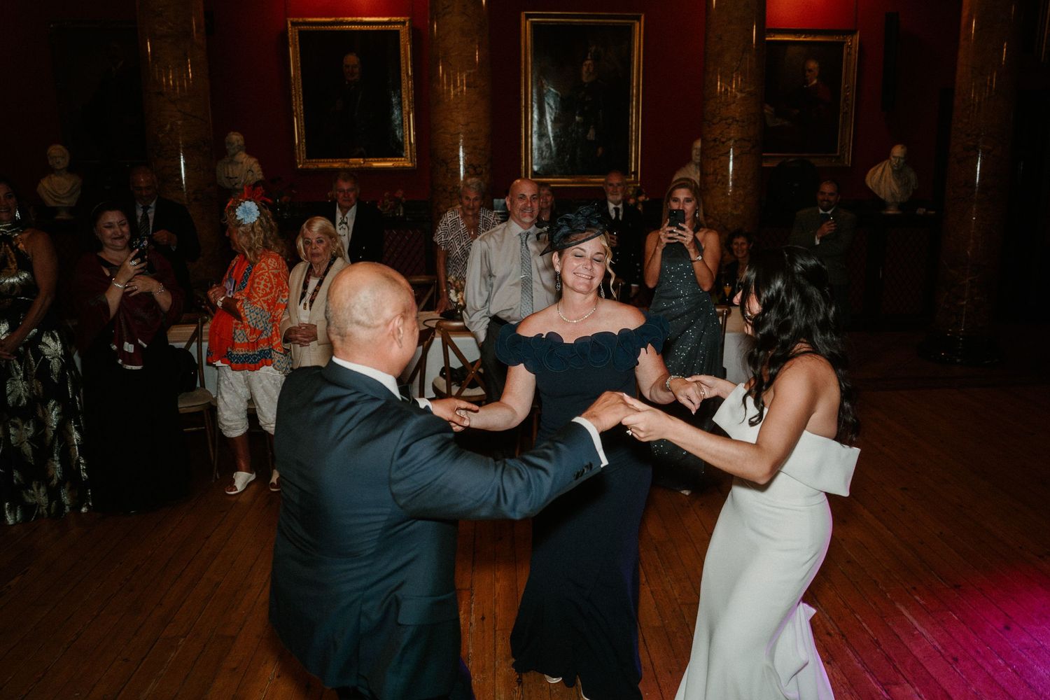 Wedding guests dancing in a historic ballroom with red walls and classical decor.