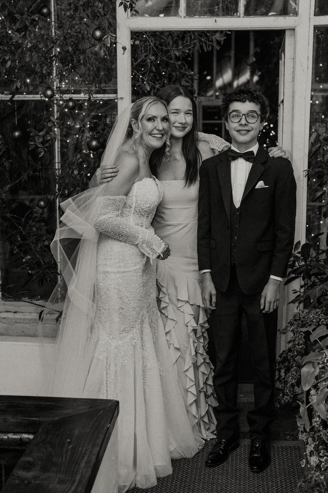 Wedding party poses together in formal attire against a backdrop of greenery and windows.
