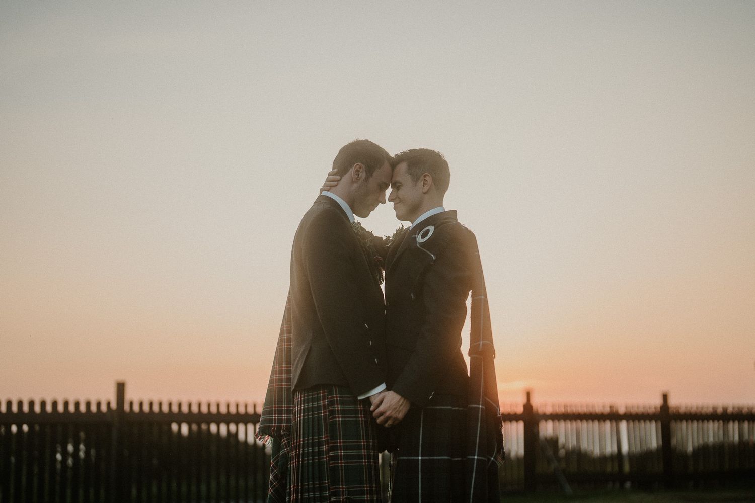 Two people in traditional Scottish dress share a tender moment at dusk.