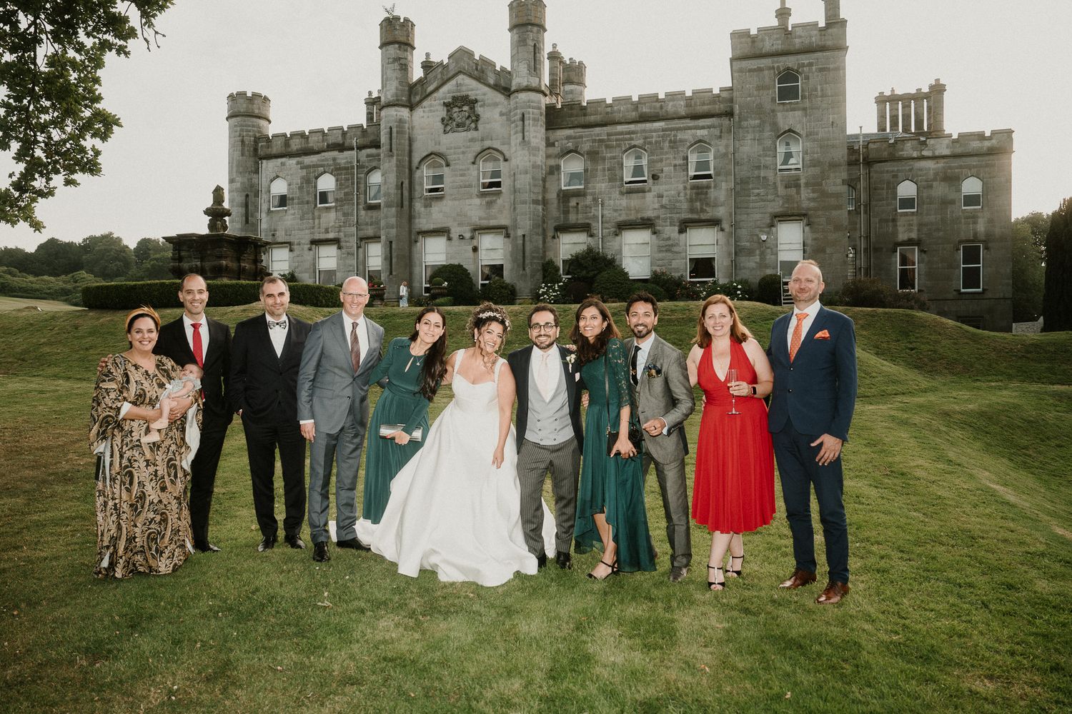 Wedding party gathered on the lawn of a grand stone castle estate for formal group photos at sunset.