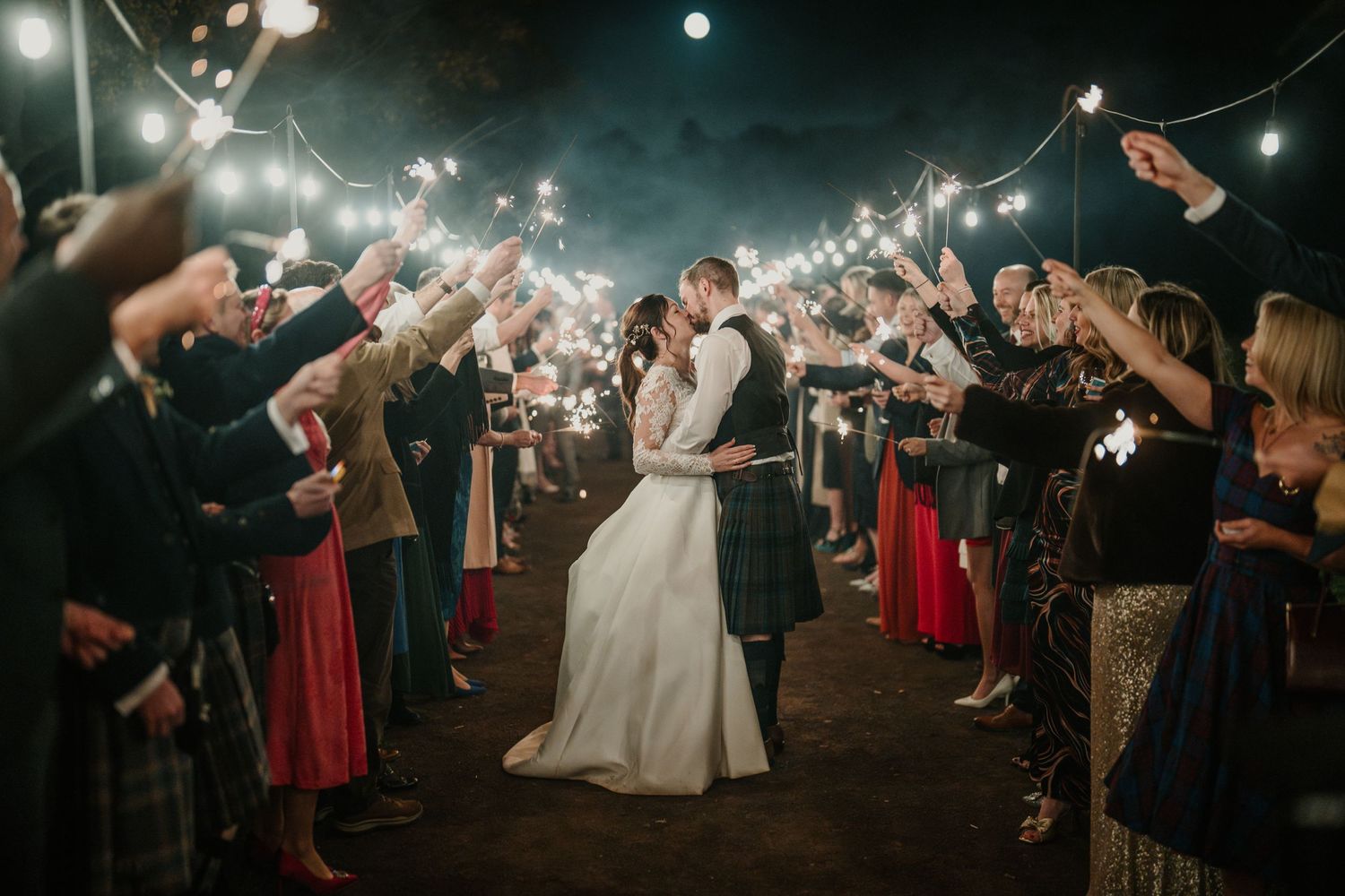 Outdoor wedding sparkler exit with guests lining pathway under string lights.