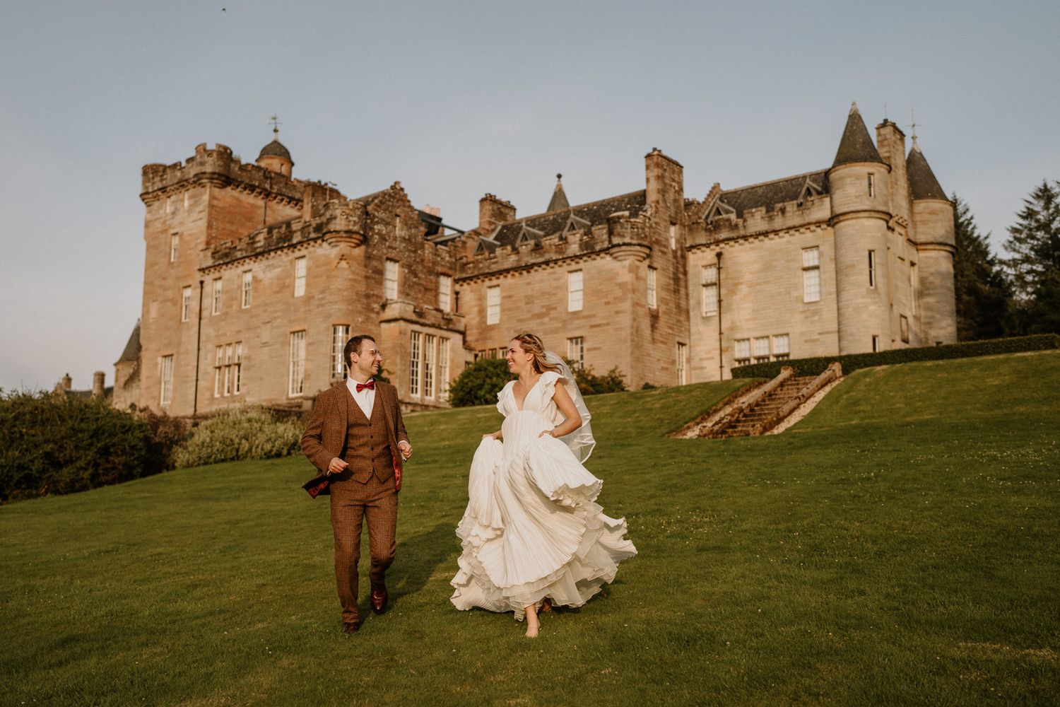 A couple walks across a green lawn in front of a majestic Scottish castle during a wedding celebration.