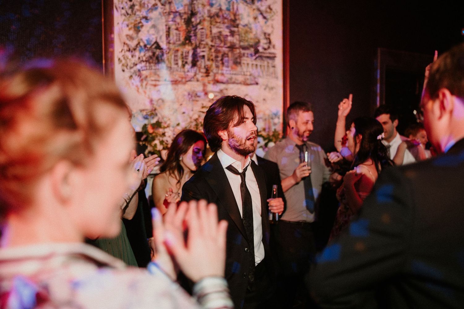 Wedding reception guests dancing with raised arms in a dimly lit celebration space.