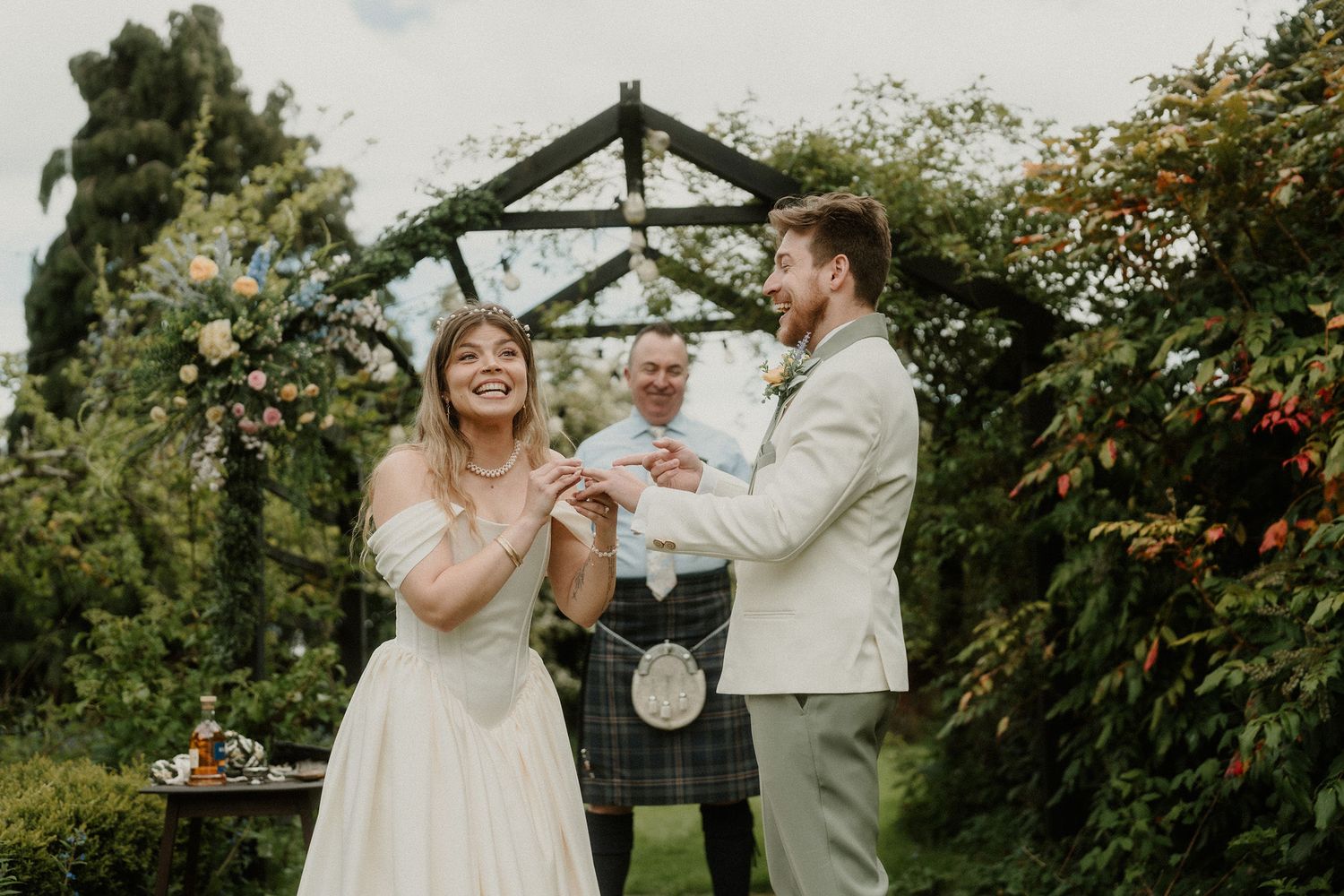 Bride and groom exchanging rings outdoors at a garden wedding ceremony with officiant in Scottish kilt.
