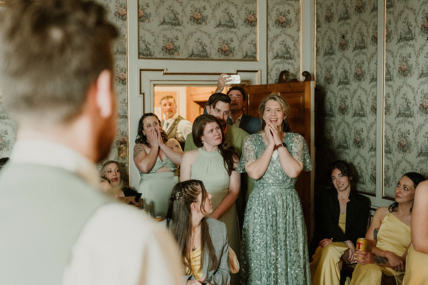 Emotional bridesmaids in sage green dresses react with joy and surprise during a wedding first look reveal indoors.