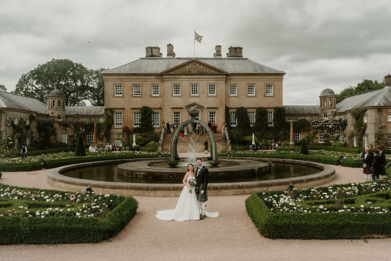 Bride and groom pose by ornate fountain in formal gardens of elegant Georgian country house wedding venue.