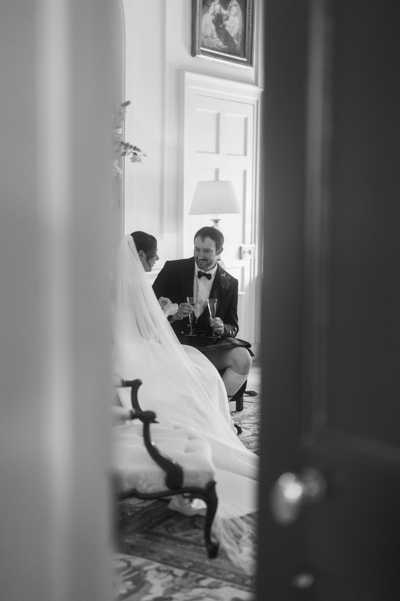 Black and white photo of bride and groom toasting champagne glasses while seated together in an elegant room.