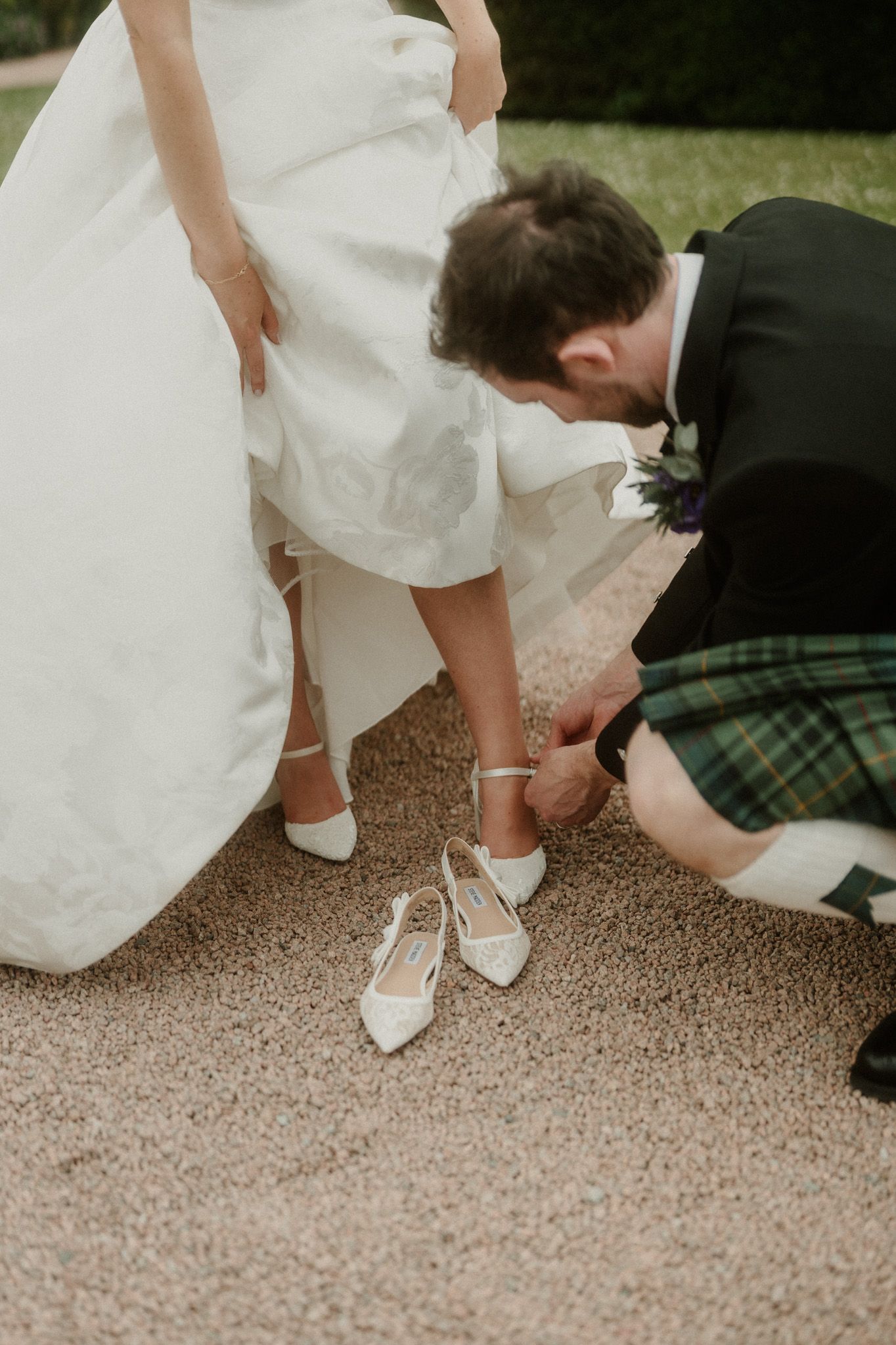 Groom in green tartan kilt fastening brides white heels on gravel path at wedding ceremony.
