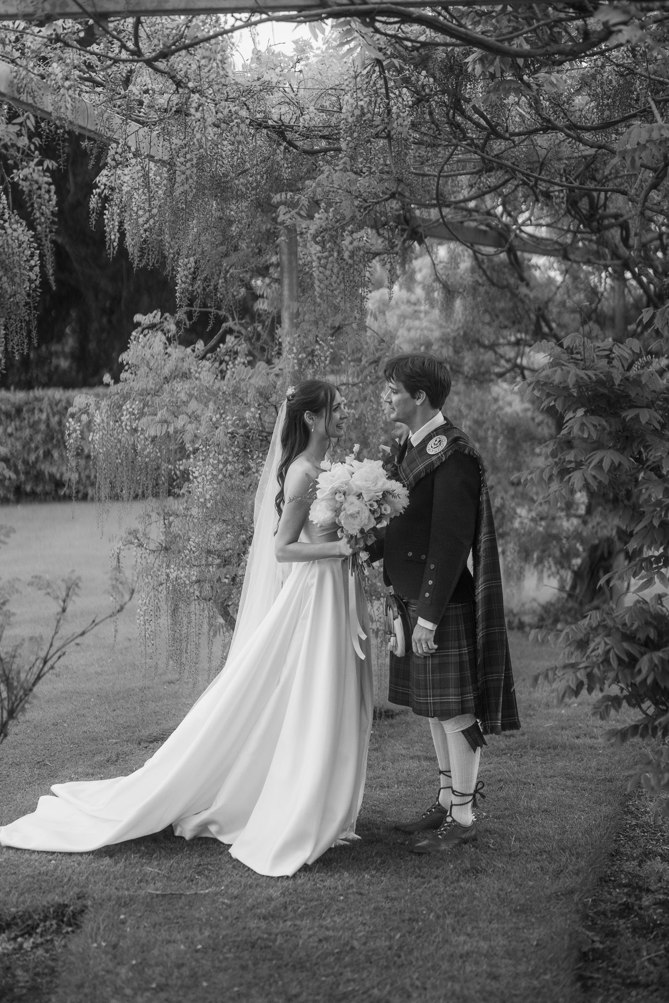 Black and white photo of bride in white gown and groom in Scottish kilt standing together in a lush garden.