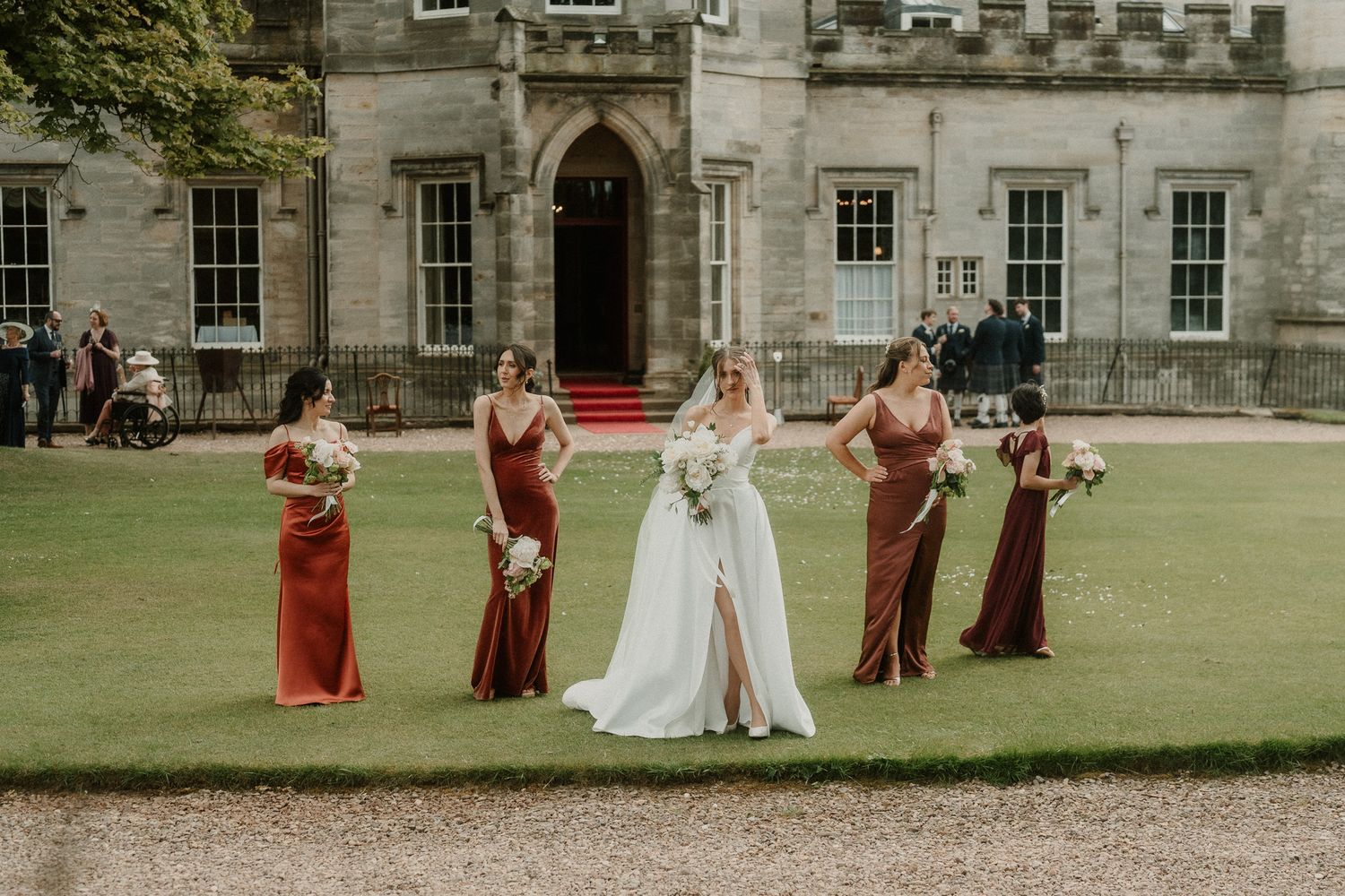 Bride in white gown with bridesmaids in red dresses posing on lawn outside elegant stone manor house.