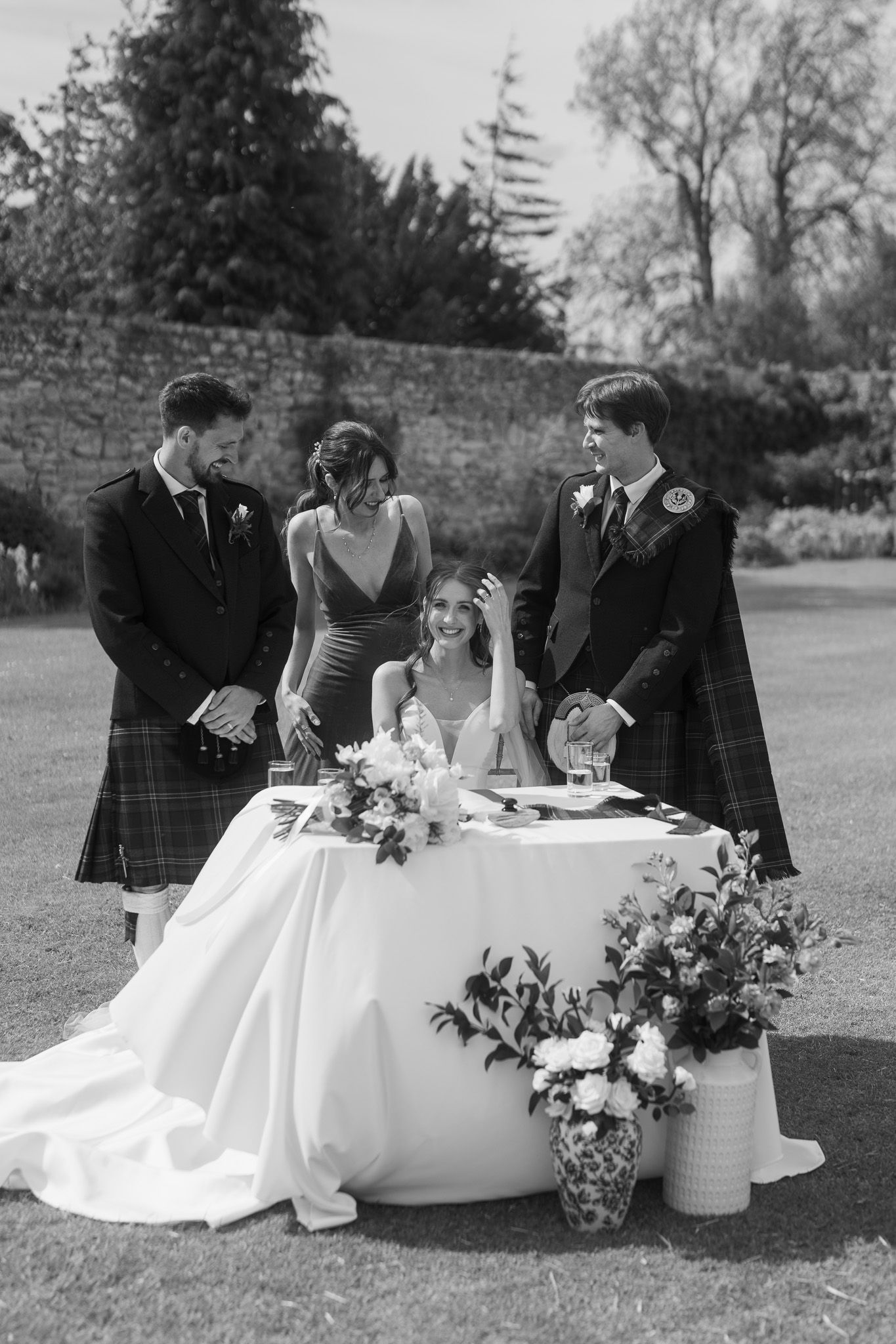 Black and white photo of a bride and groom signing wedding documents outdoors, flanked by guests in Scottish kilts.