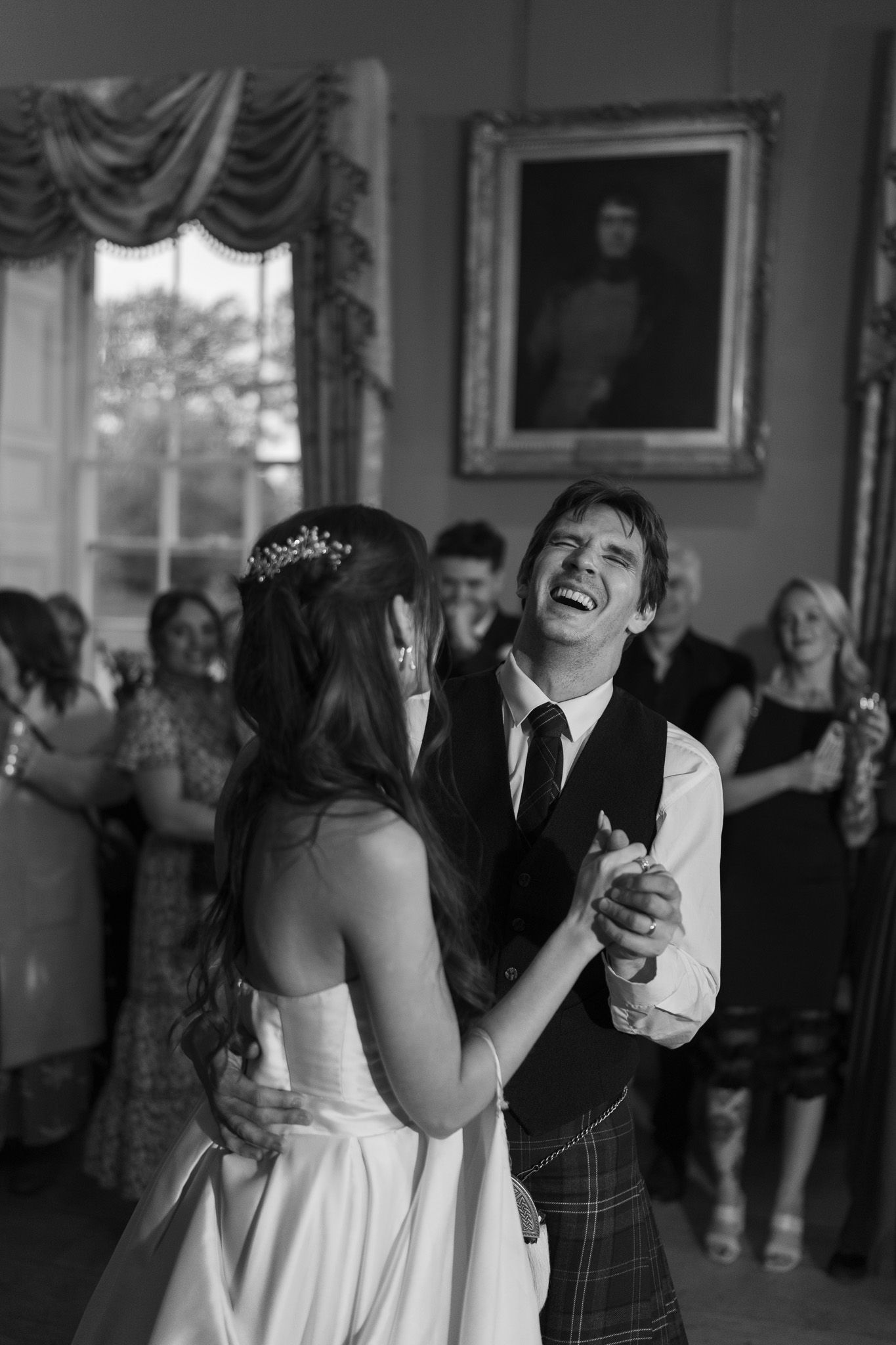 Black and white photo of a joyful groom in a kilt sharing a first dance with his bride at a wedding reception.