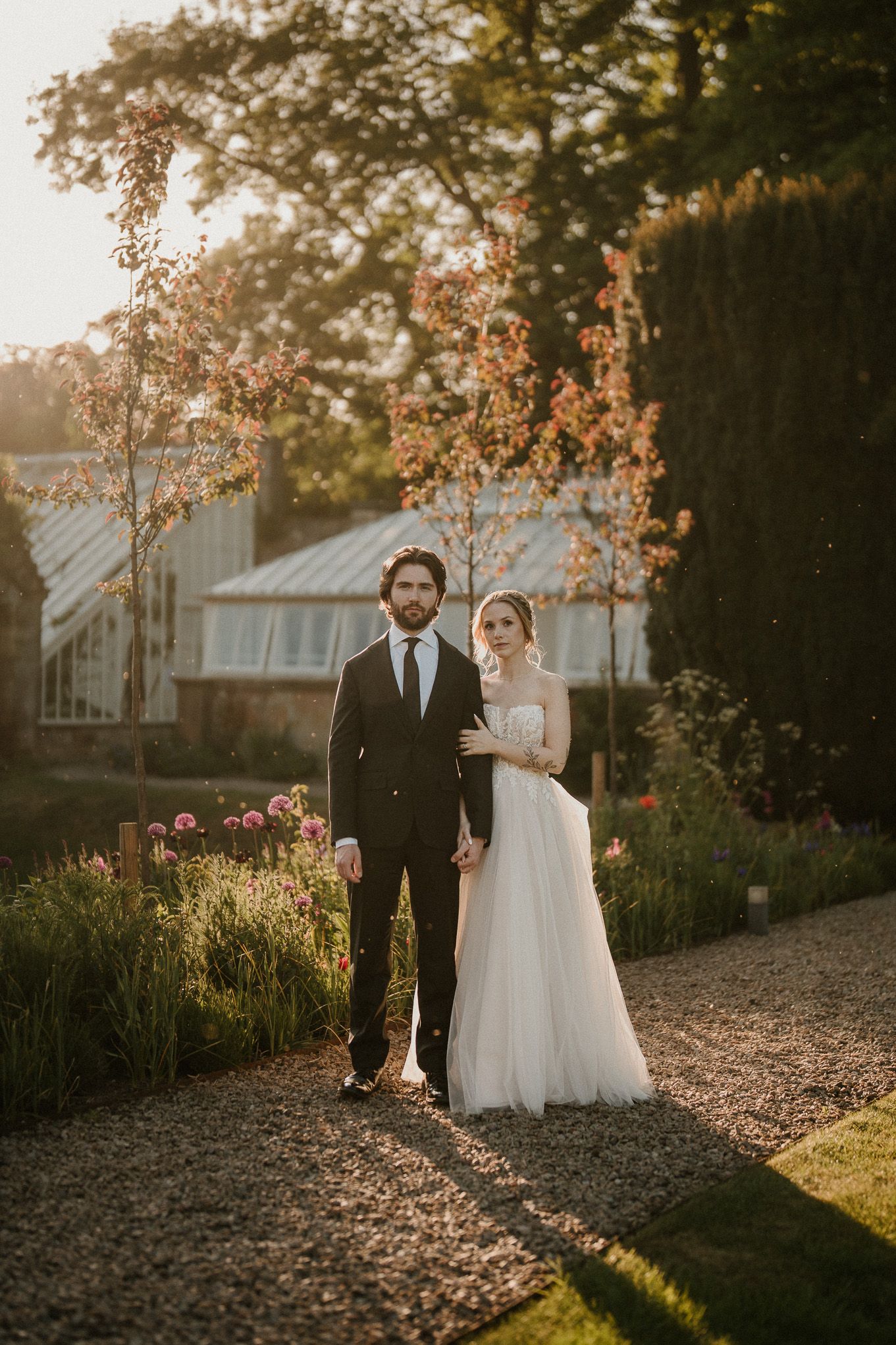 Bride and groom stand on a garden path at golden hour, surrounded by blooming flowers and lush greenery.