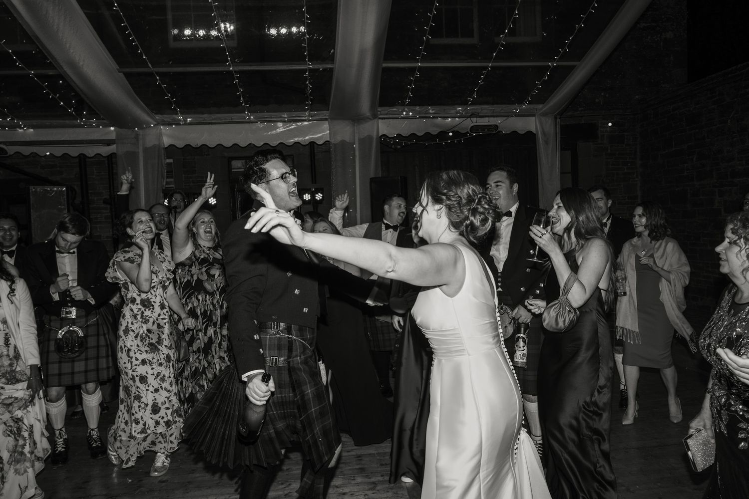 Black and white photo of a groom in a kilt dancing with a bride in a long gown as wedding guests cheer and dance behind them.