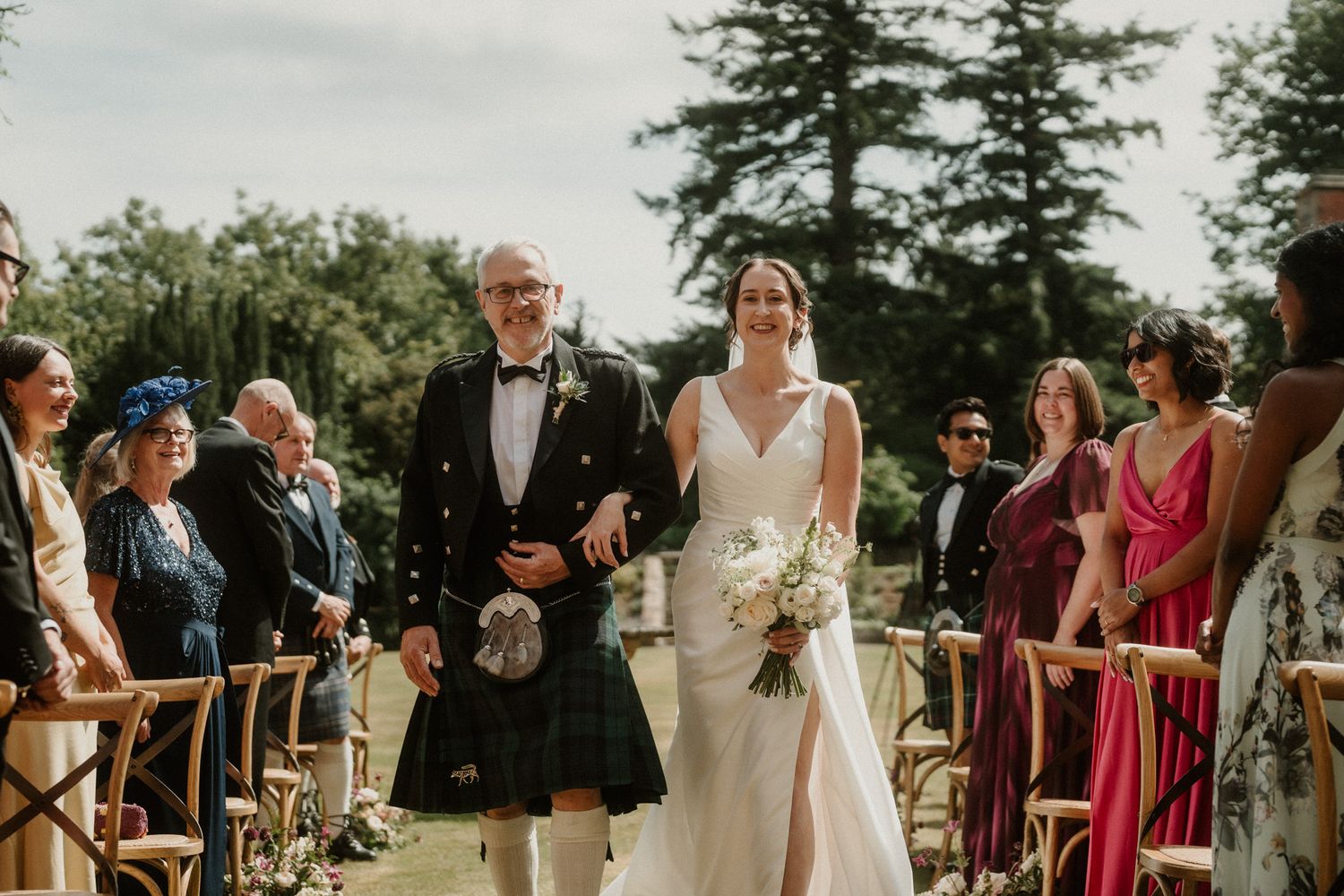 Bride in a white gown carrying a bouquet walks down the aisle on the arm of a kilted man as outdoor wedding guests look on.