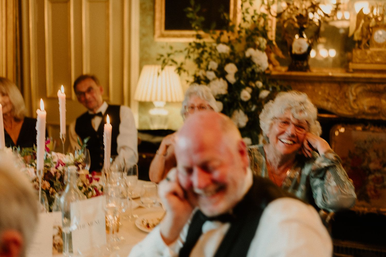 Guests laugh and smile at an elegant candlelit wedding reception table with white flowers, wine glasses and warm ambient lighting.