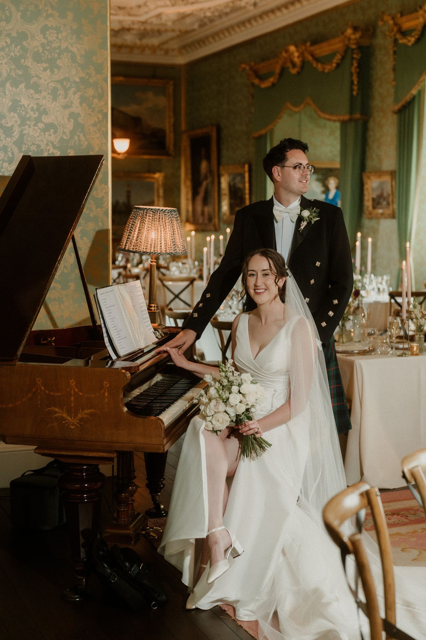 Bride in a white gown and veil holding a bouquet sits beside a groom in a kilt jacket at a grand piano in an elegant wedding venue.