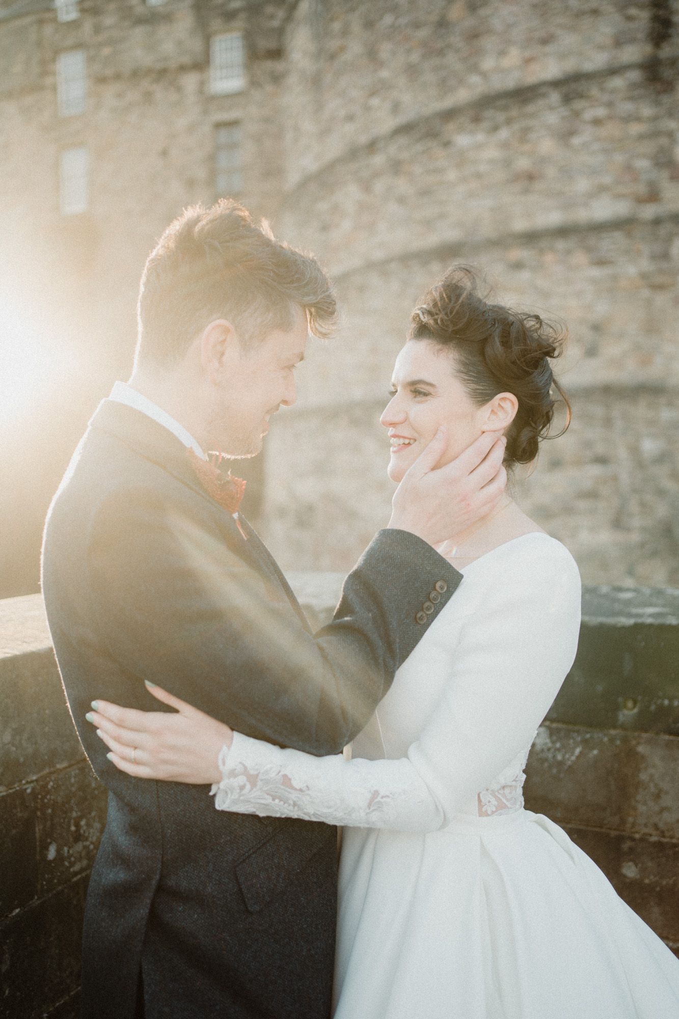 Tender moment between newlyweds captured in warm sunlight against stone castle wall.