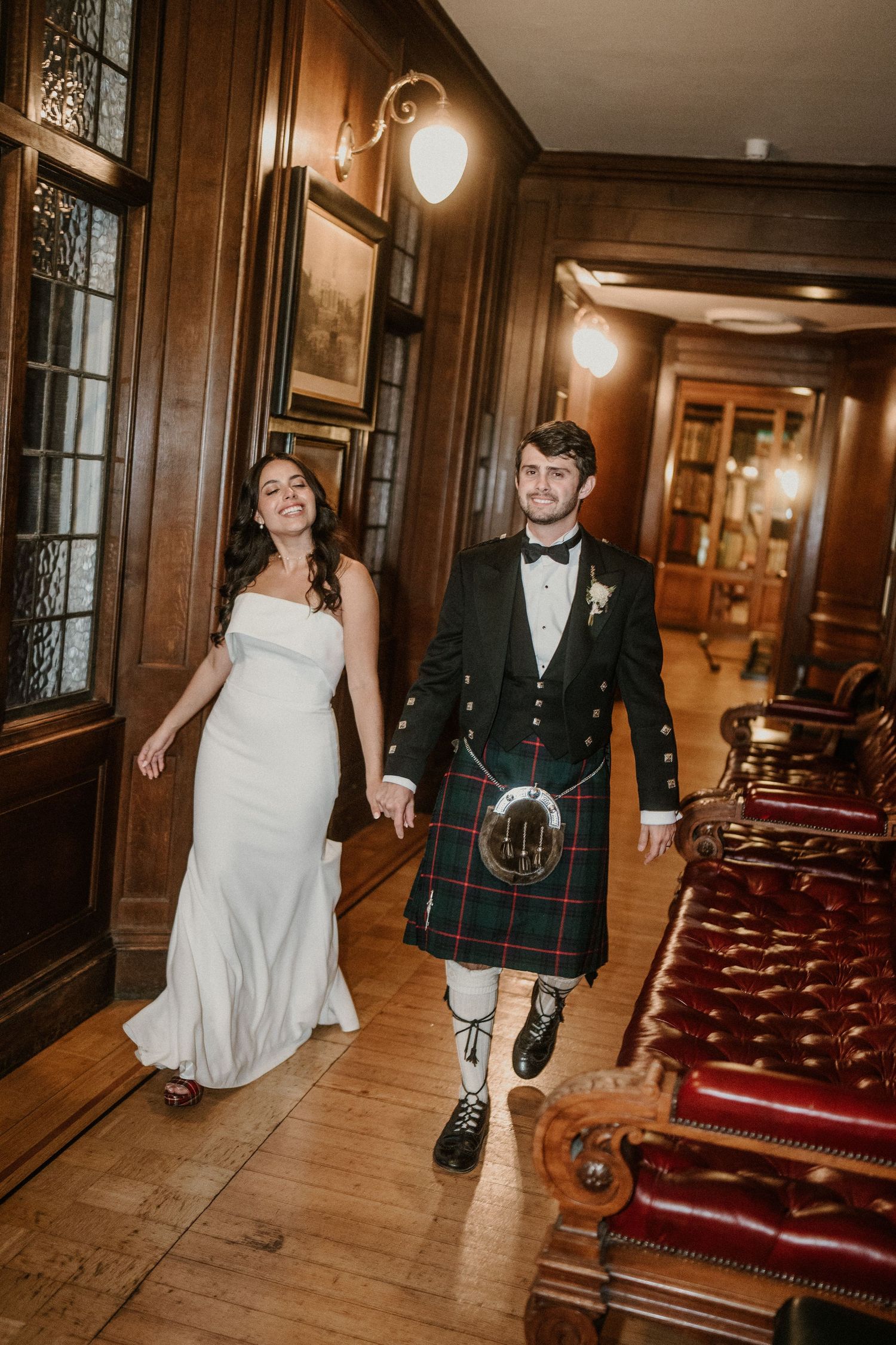 Couple walking through a wood-paneled hallway with Scottish wedding attire.