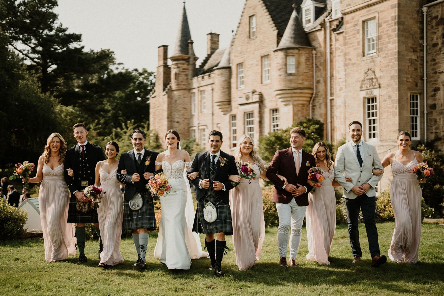 Wedding party walking and laughing together on lawn in front of stone mansion.