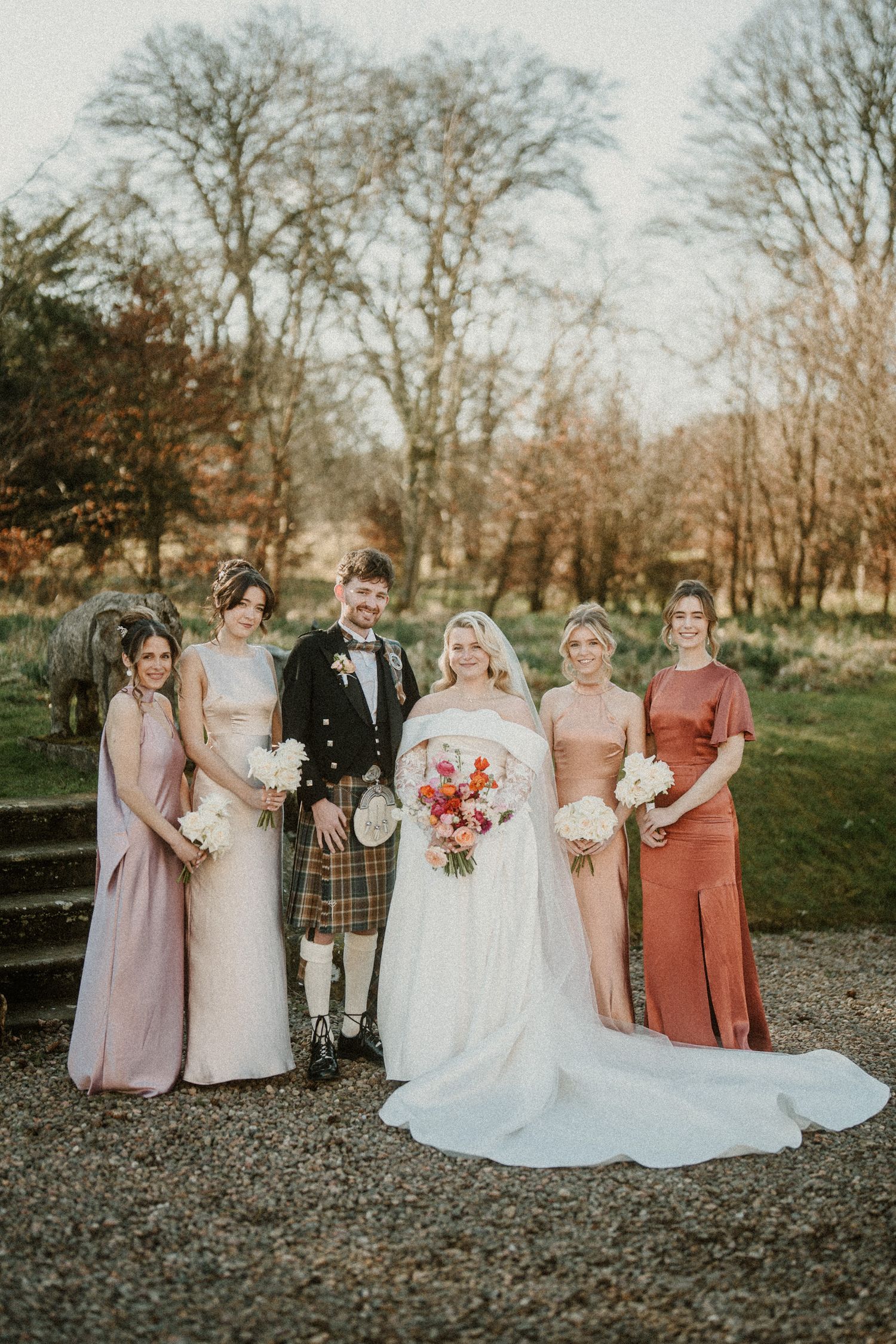Wedding party in pastel and coral dresses pose against autumn trees.