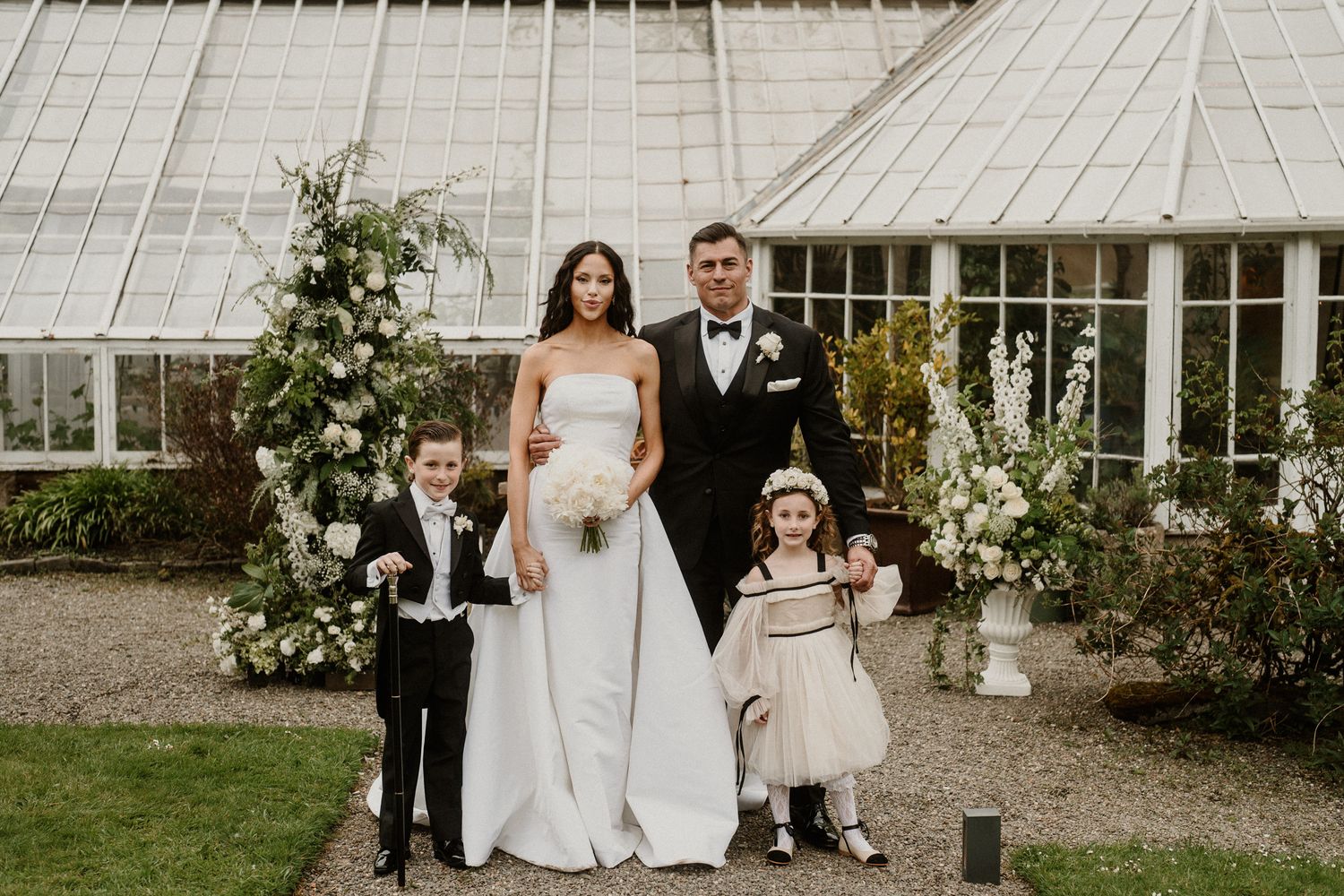 Wedding party stands together in front of glass greenhouse with white floral decor.