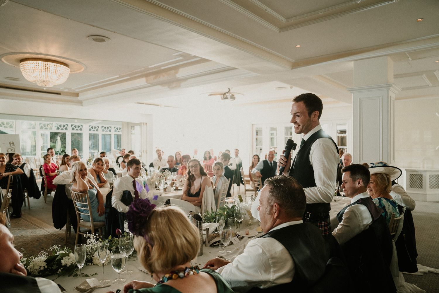 Wedding celebration in a bright ballroom with guests seated at decorated tables.