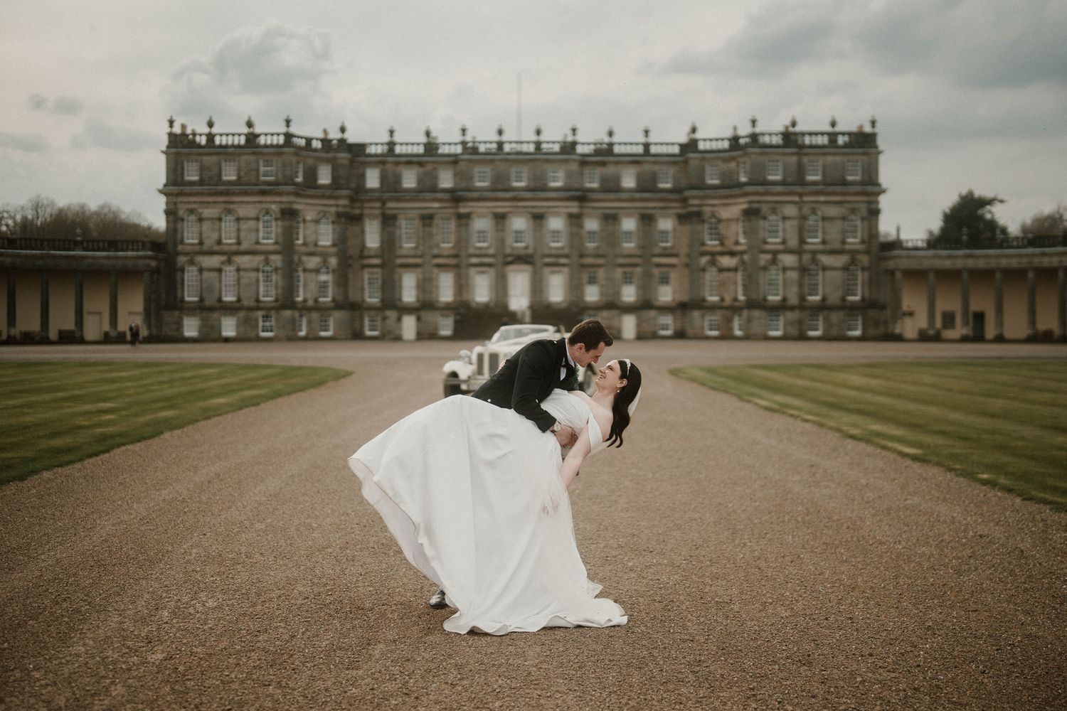A romantic couple shares a kiss on the grand driveway in front of a historic English manor house on their wedding day.