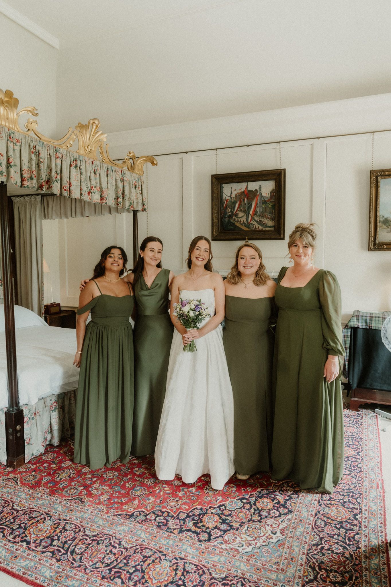 Bride in white gown holds bouquet flanked by three bridesmaids in olive green dresses inside an elegant bedroom.