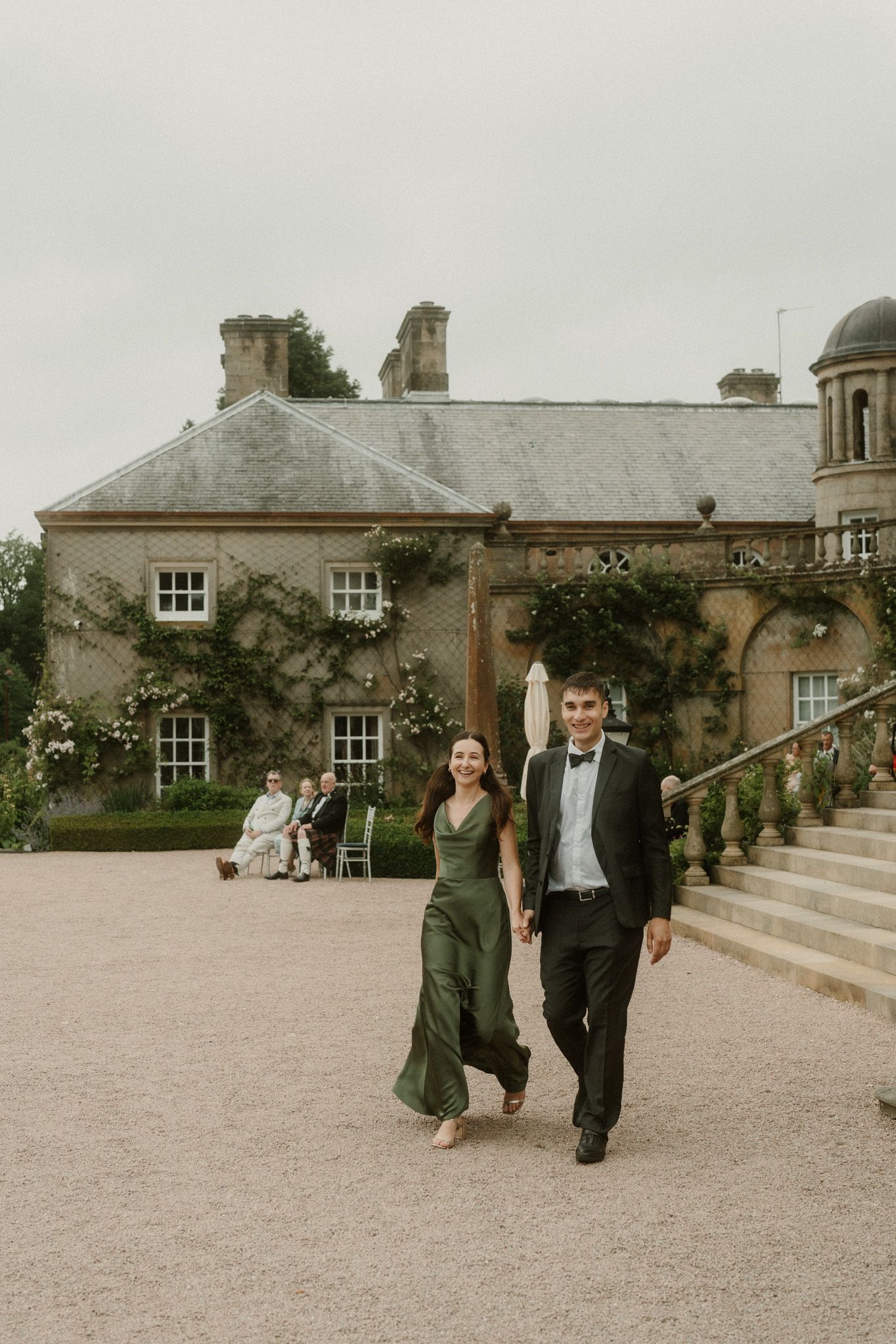 Couple in formal attire walking hand-in-hand outside an elegant ivy-covered stone manor house.
