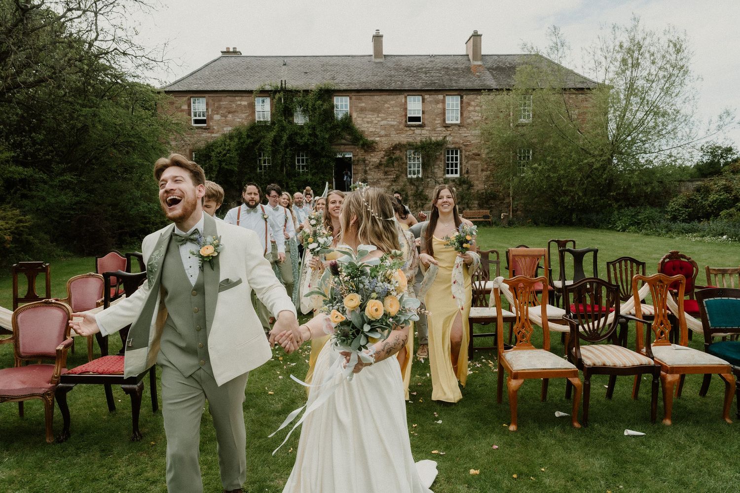 Joyful bride and groom walk hand-in-hand after outdoor wedding ceremony, with guests and mismatched chairs on green lawn.